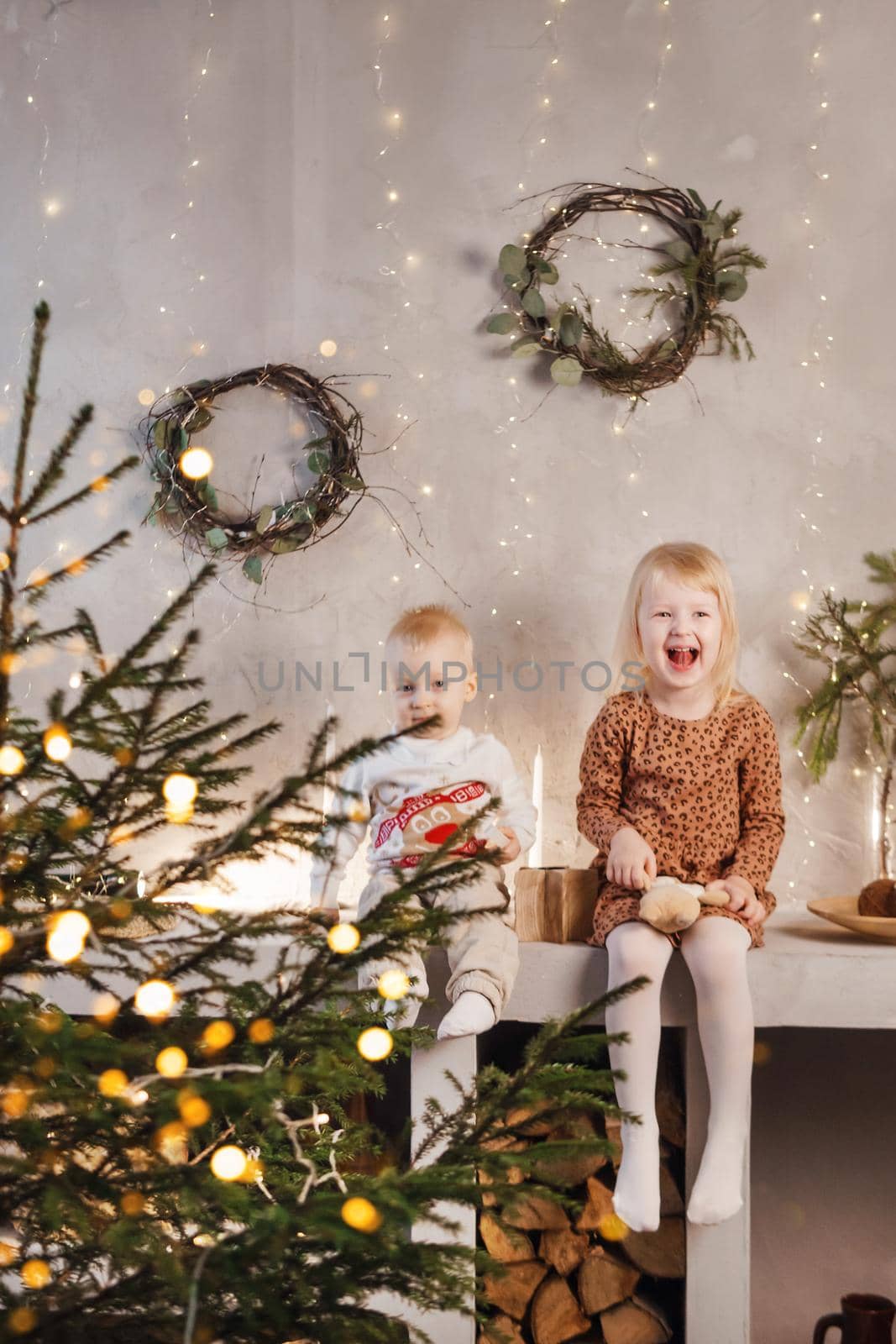 Little brother and sister play on Christmas eve in a beautiful house decorated for the New Year holidays. Children are playing with a Christmas gift. Scandinavian-style interior with live fir trees and a wooden staircase.