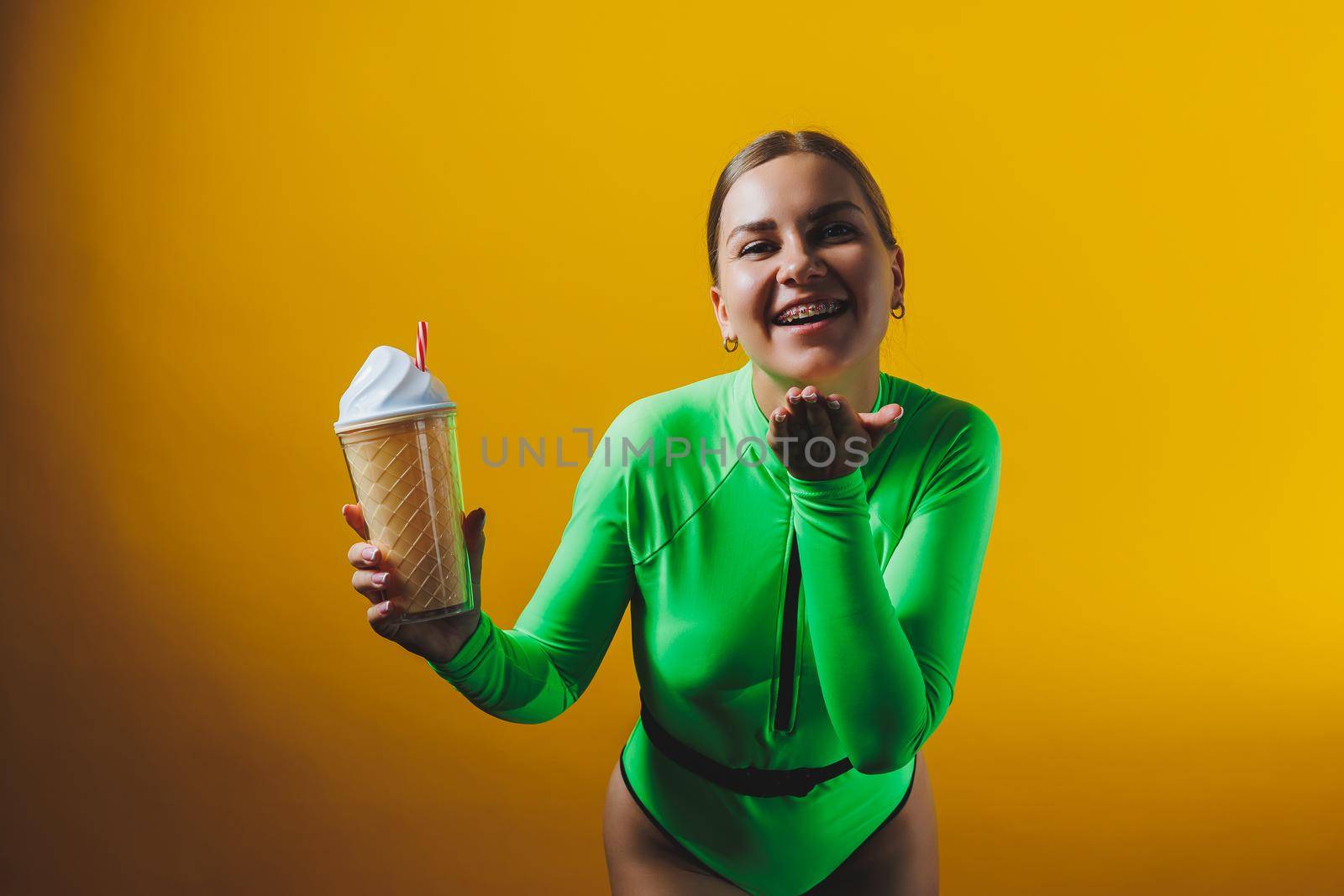 A smiling woman in a green swimsuit with a cocktail in her hands is happy about the upcoming vacation, isolated on a bright yellow background. Summer tropics travel concept.