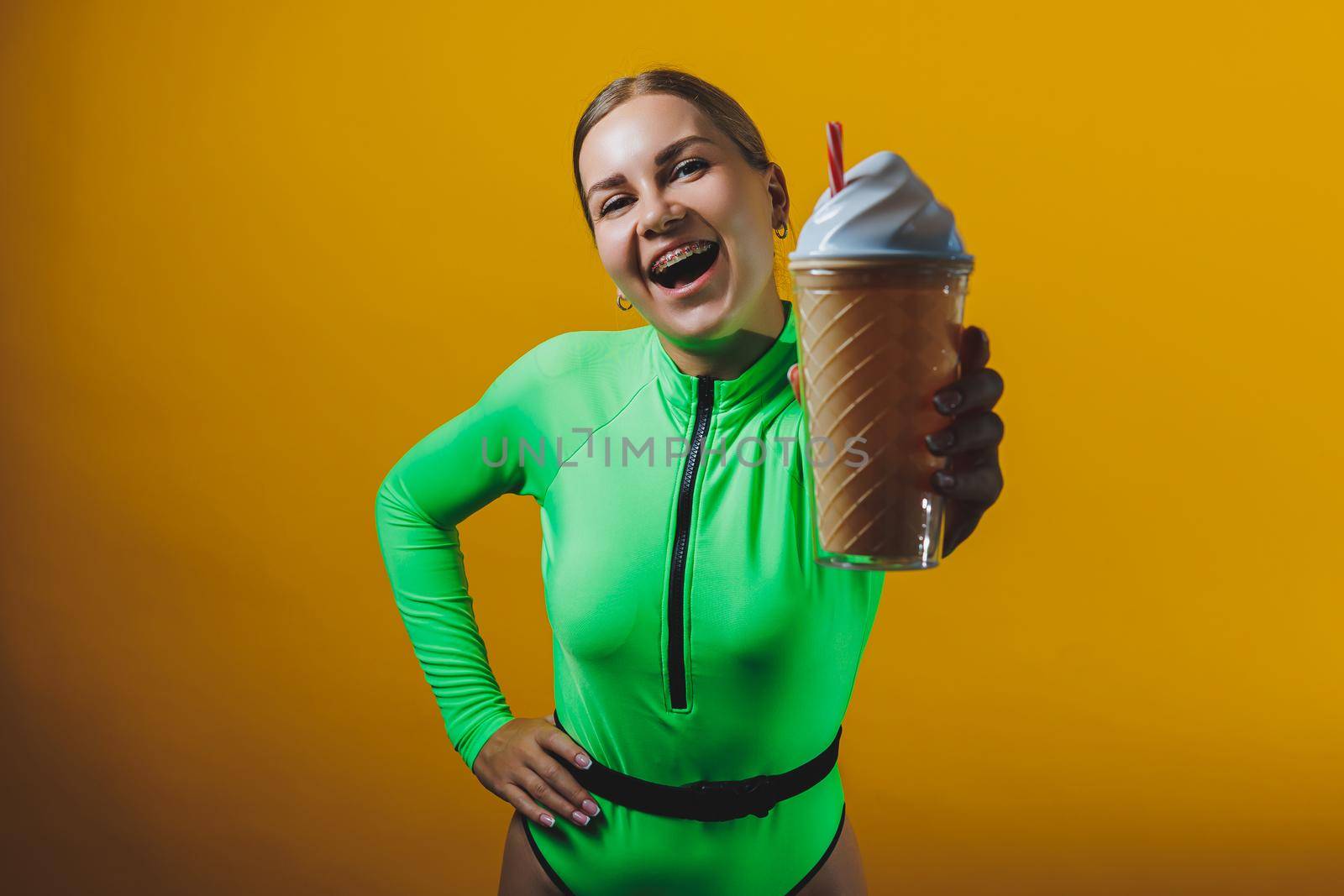 A young smiling blonde in a green swimsuit with a cocktail in her hands is happy about the upcoming vacation, isolated on bright yellow. Summer tropics travel concept.