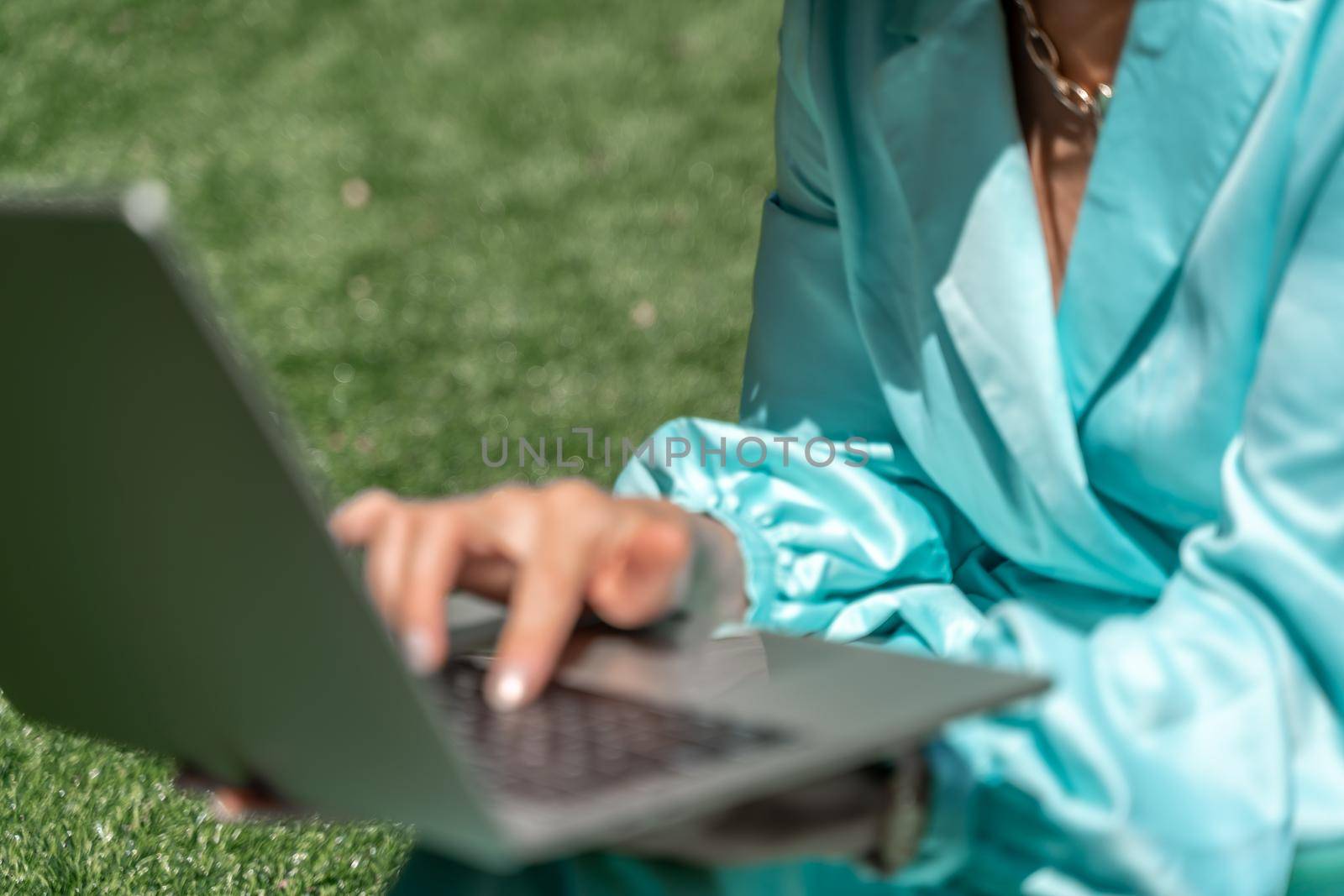 a young beautiful woman with blond curly hair in glasses and a blue dress sits on the grass in nature and uses a laptop