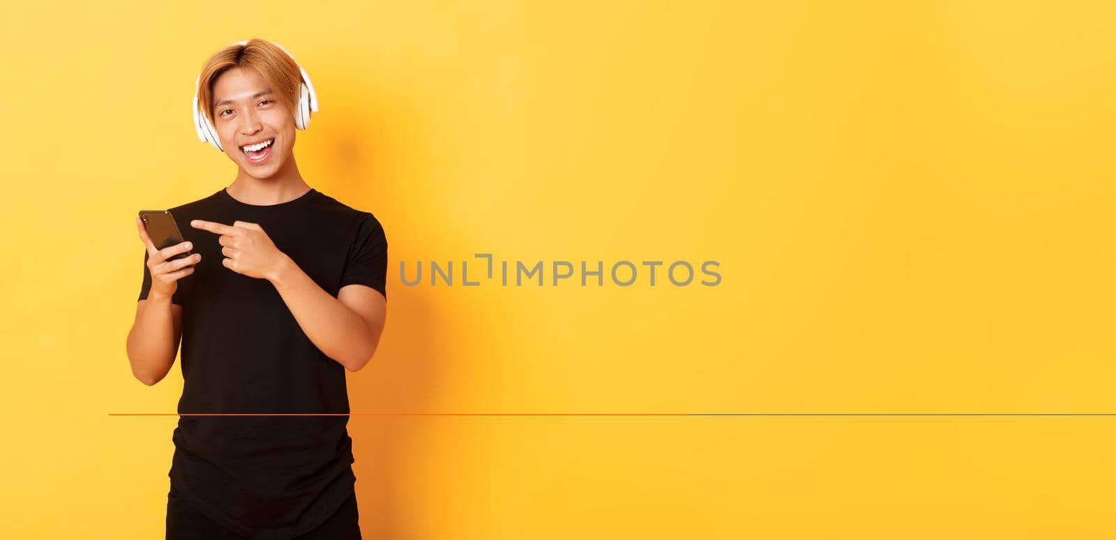 Joyful satisfied asian handsome guy, listening music or good podcast in headphones, pointing finger at smartphone with pleased smile, yellow background by Benzoix
