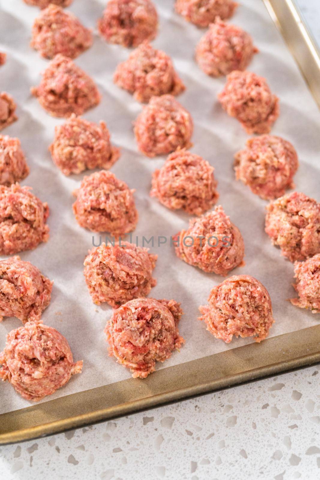 Scooping ground meat with dough scoop into a baking sheet lined with parchment paper to prepare oven-baked meatballs.