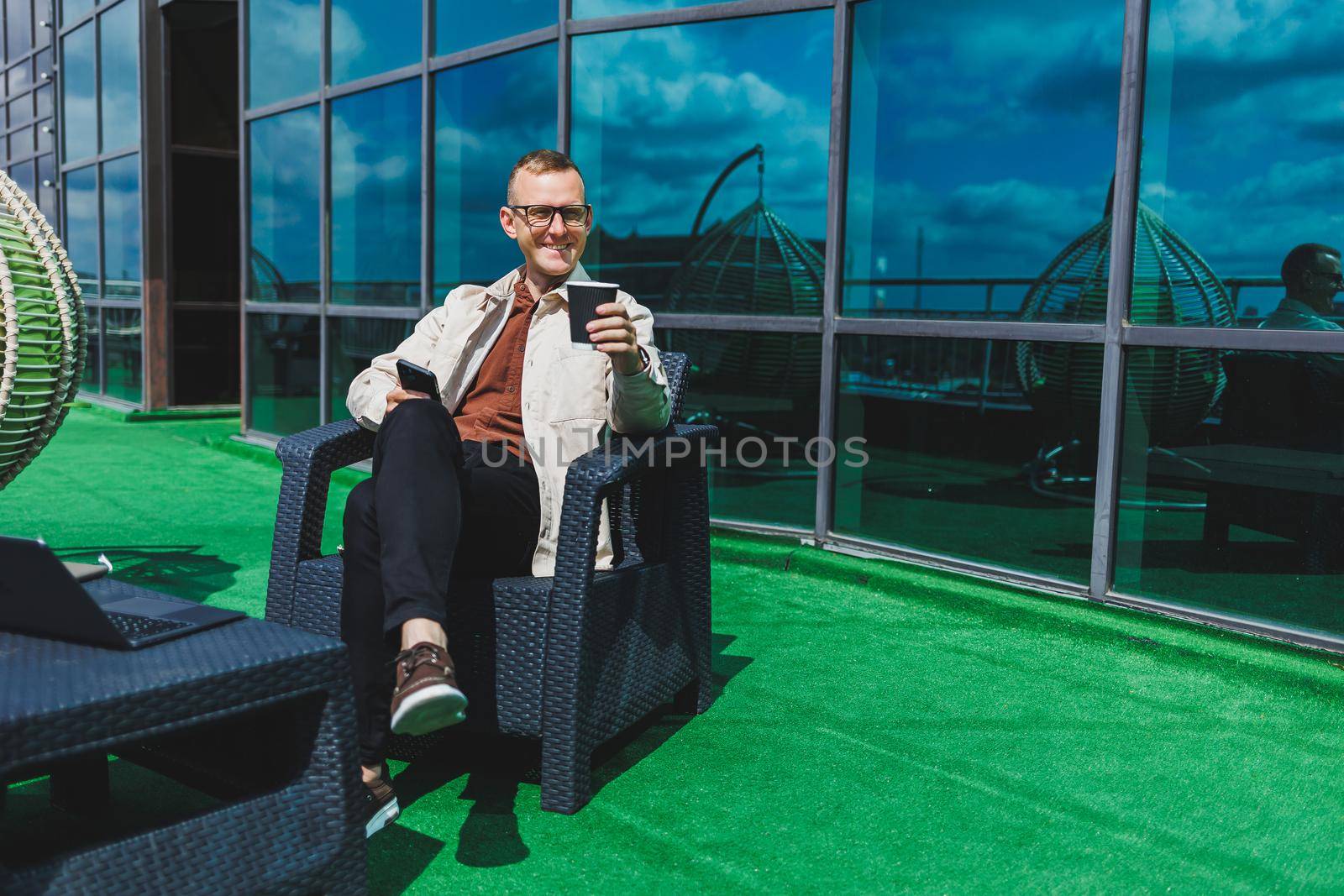 Confident young businessman, dressed in formal wear and stylish glasses, calls on a smartphone while sitting with coffee, on the terrace of a modern office building with a glass wall