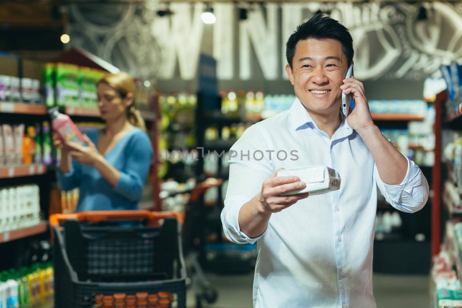 A young handsome Asian man stands at the department of household chemicals, chooses pads in the supermarket . He calls his wife, girlfriend, consults, asks, smiles.