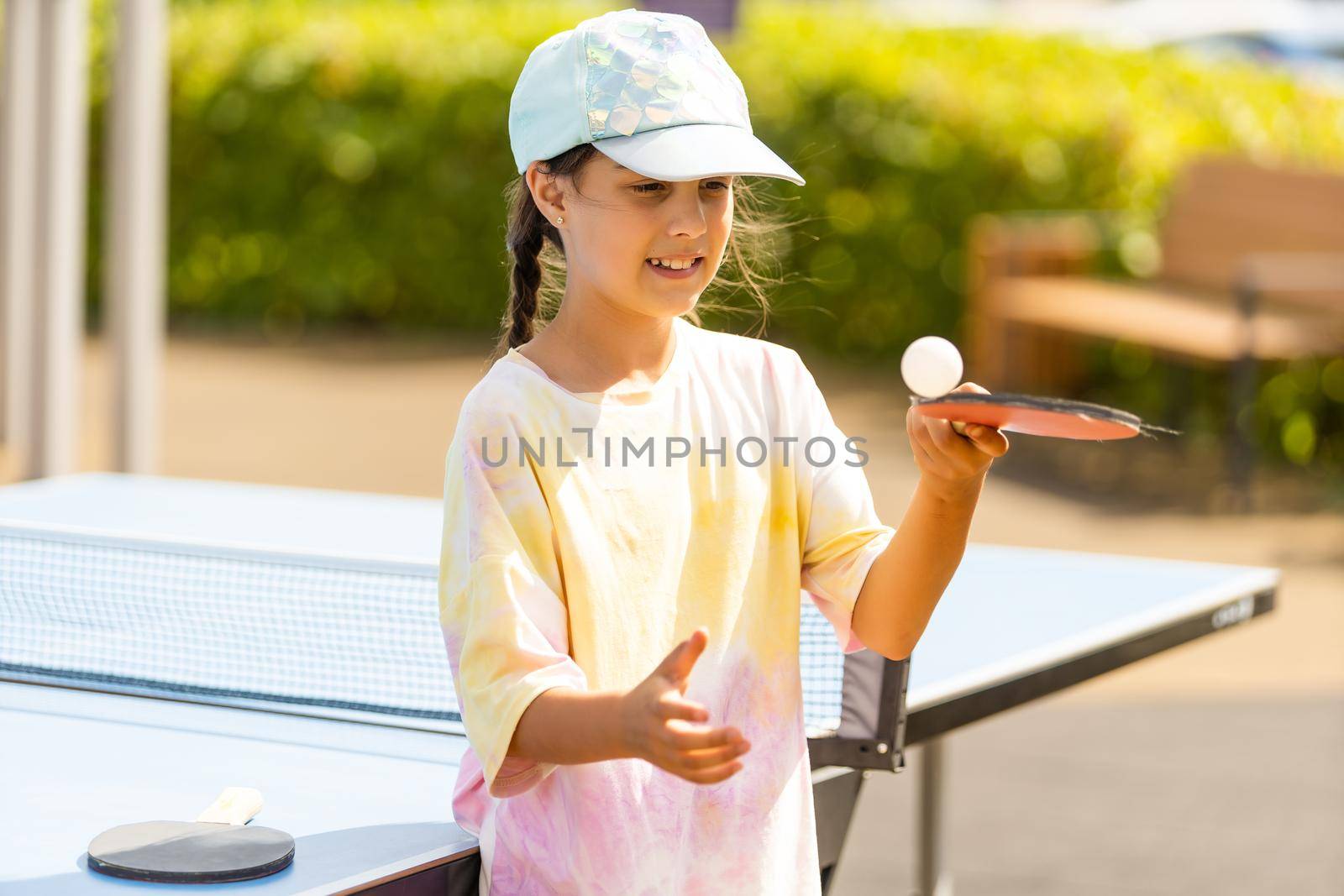 Young teenager girl playing ping pong. She holds a ball and a racket in her hands. Playing table tennis outdoors in the yard. by Andelov13