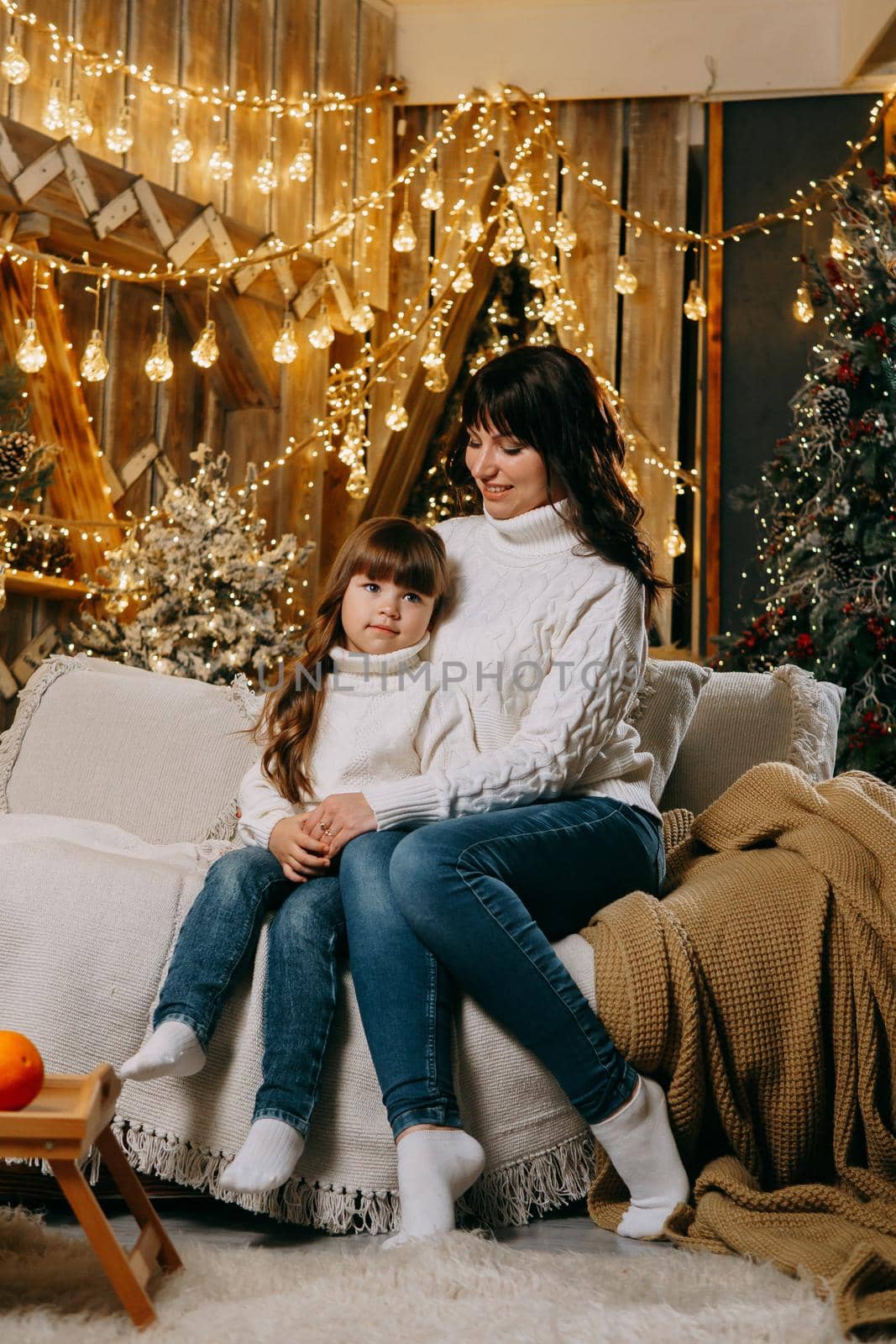 A little girl with her mother in a cozy home environment on the sofa next to the Christmas tree. The theme of New Year holidays and festive interior with garlands and light bulbs.