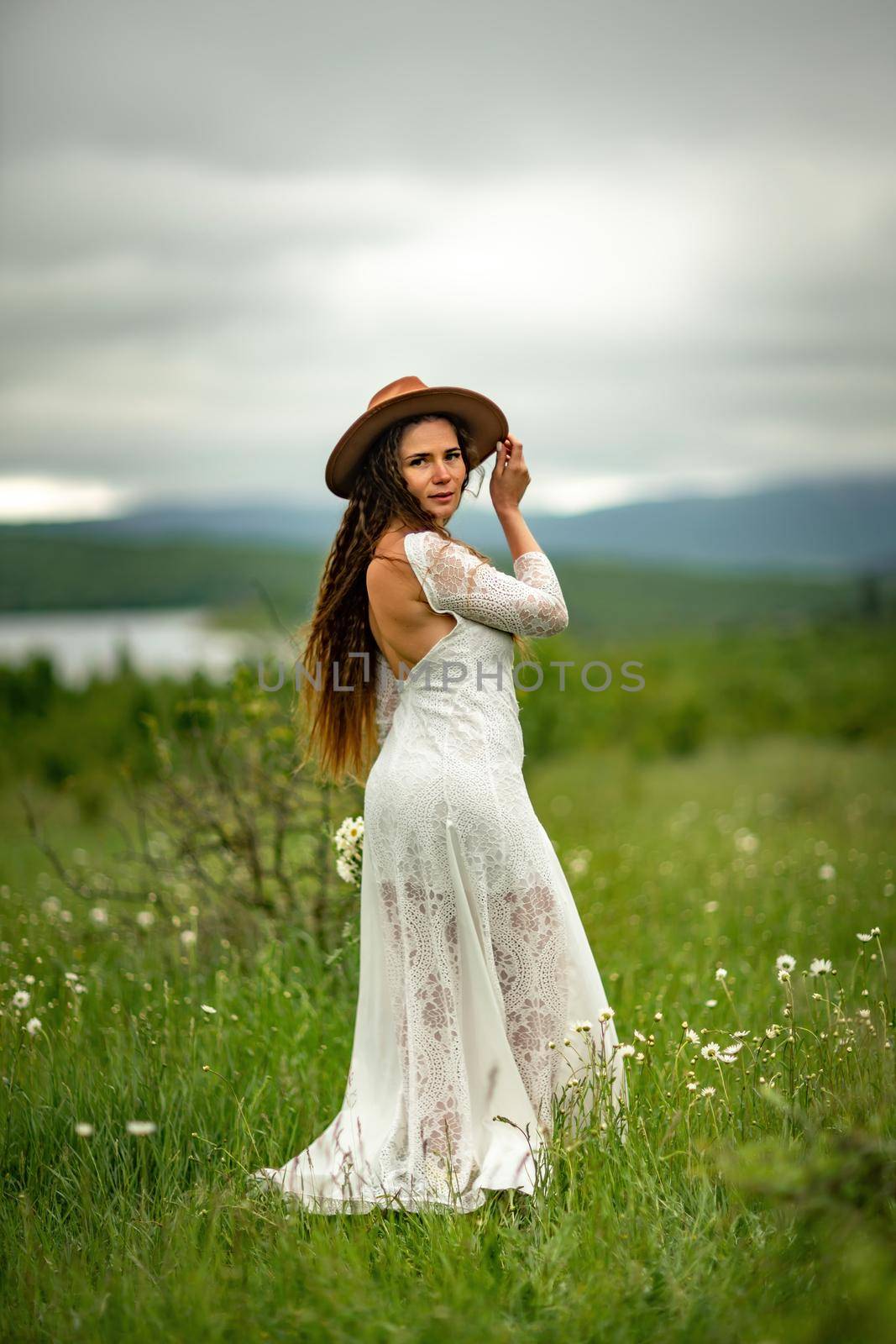 A middle-aged woman in a white dress and brown hat stands on a green field and holds a basket in her hands with a large bouquet of daisies. In the background there are mountains and a lake