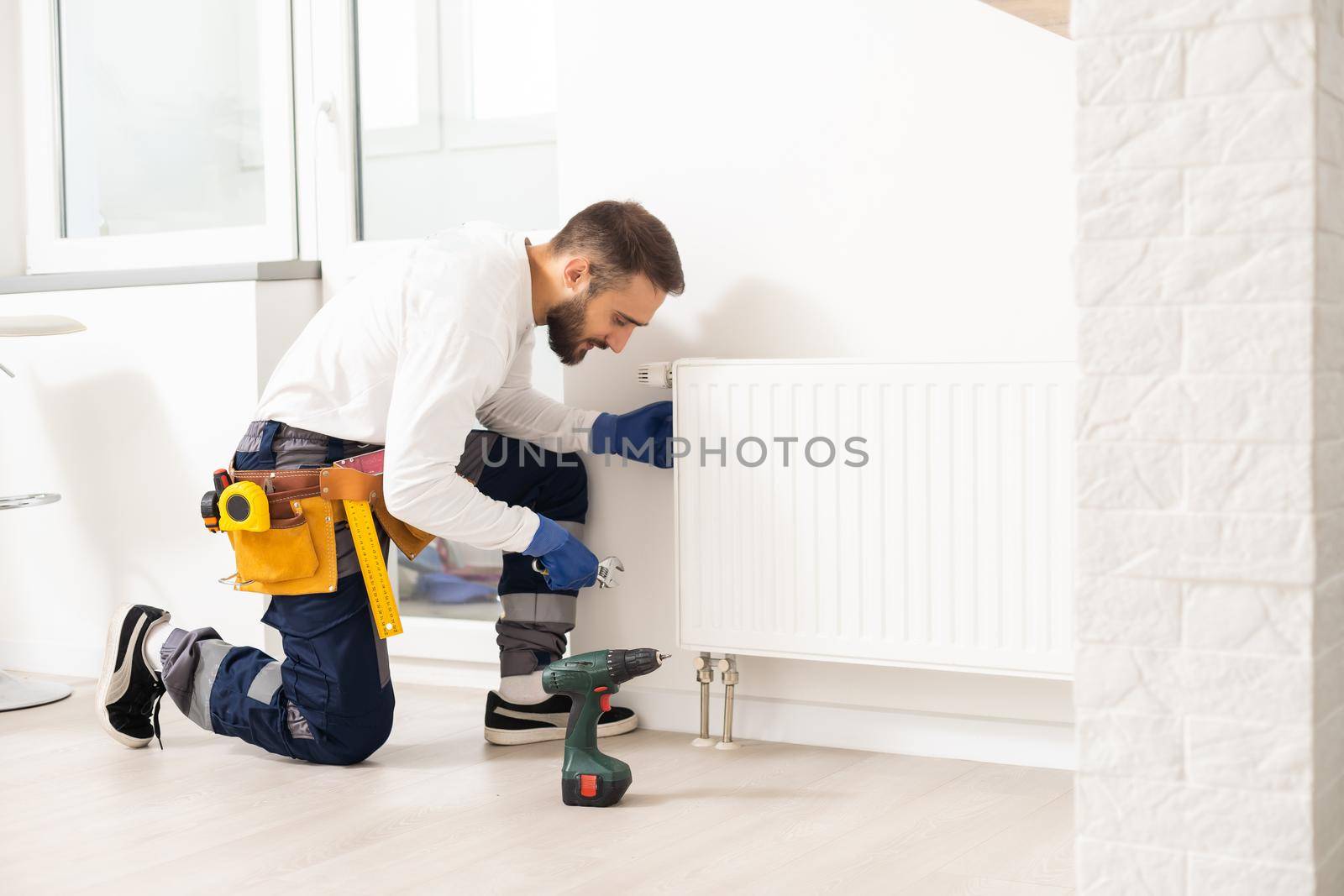 Man in work overalls using wrench while installing heating radiator in room. Young plumber installing heating system in apartment. Concept of radiator installation, plumbing works and home renovation