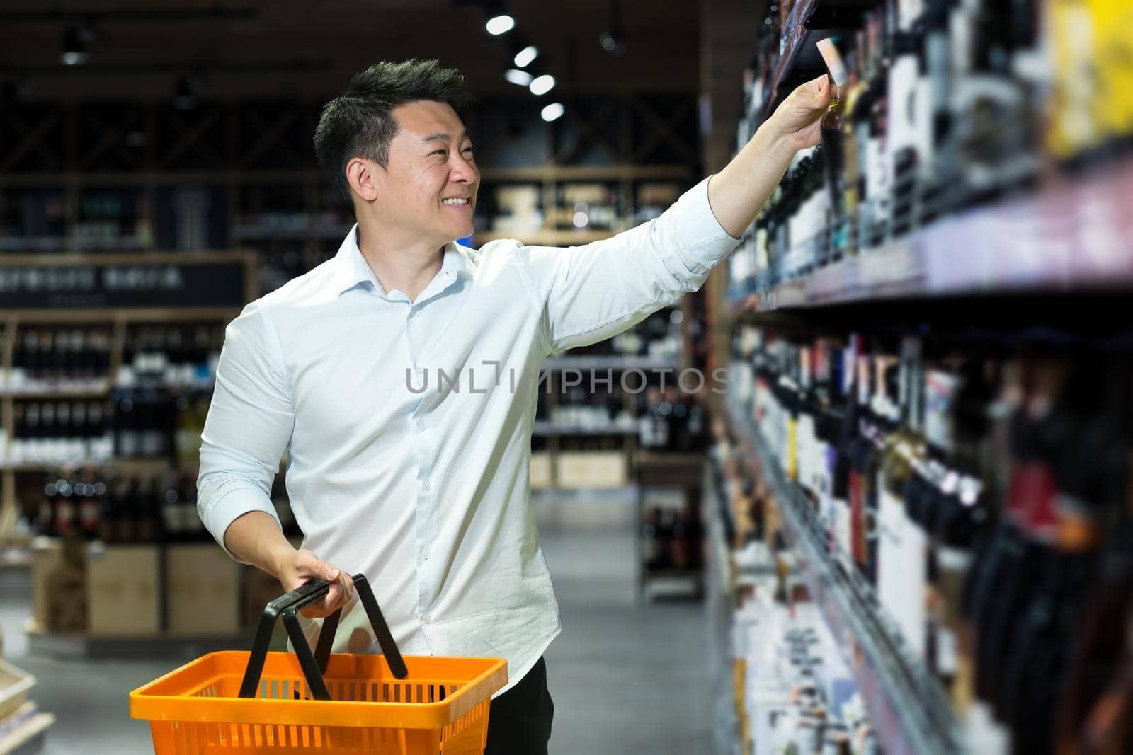 Young handsome male Asian businessman in a white shirt chooses wine in the alcohol department in a supermarket. He is holding a basket for the product in his hand, smiling.