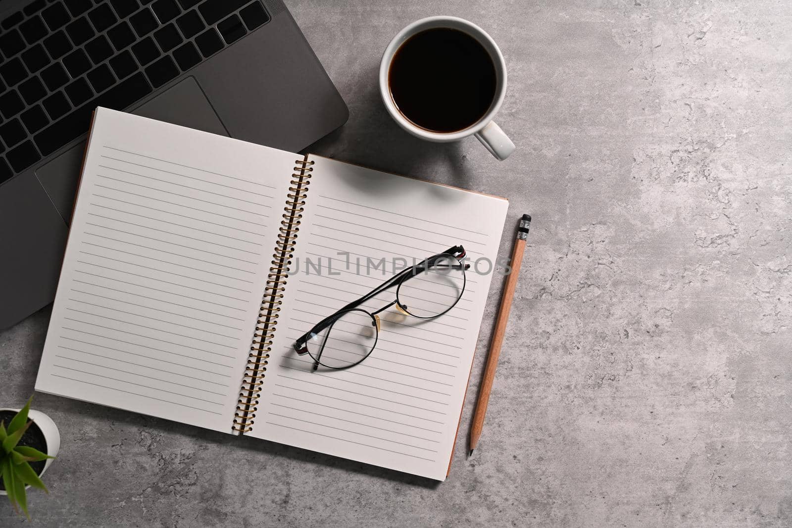 Flat lay blank notebook, eyeglasses, coffee cup and laptop on stone table.