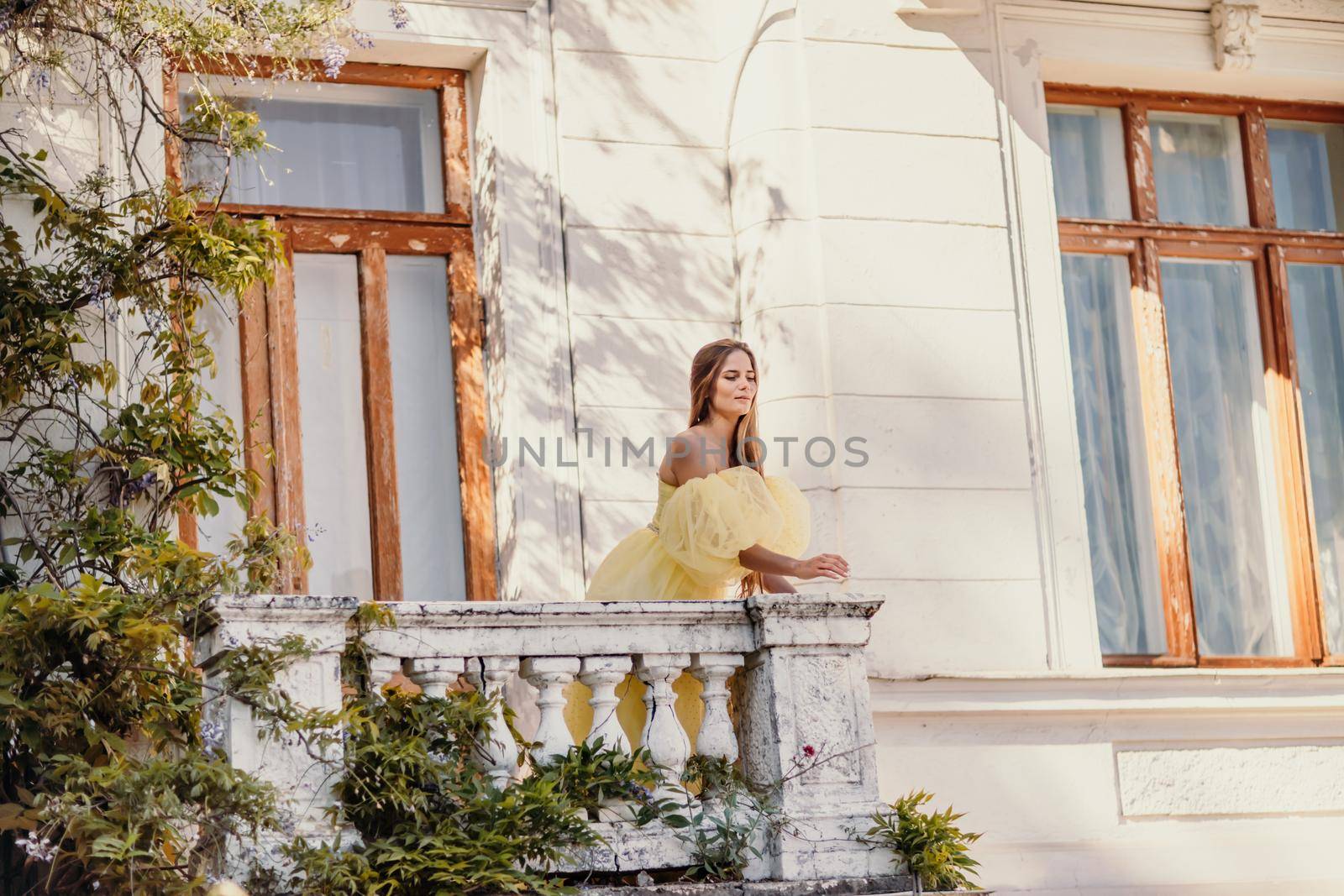 a beautiful smiling and kind woman in a gorgeous yellow dress stands on the balcony of an old vintage house.