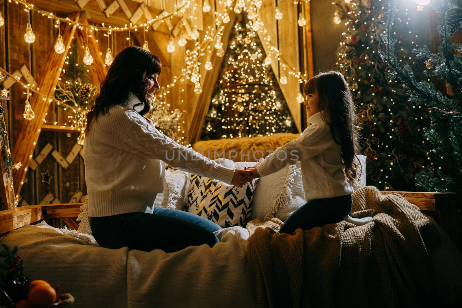 A little girl with her mother in a cozy home environment on the sofa next to the Christmas tree. The theme of New Year holidays and festive interior with garlands and light bulbs.