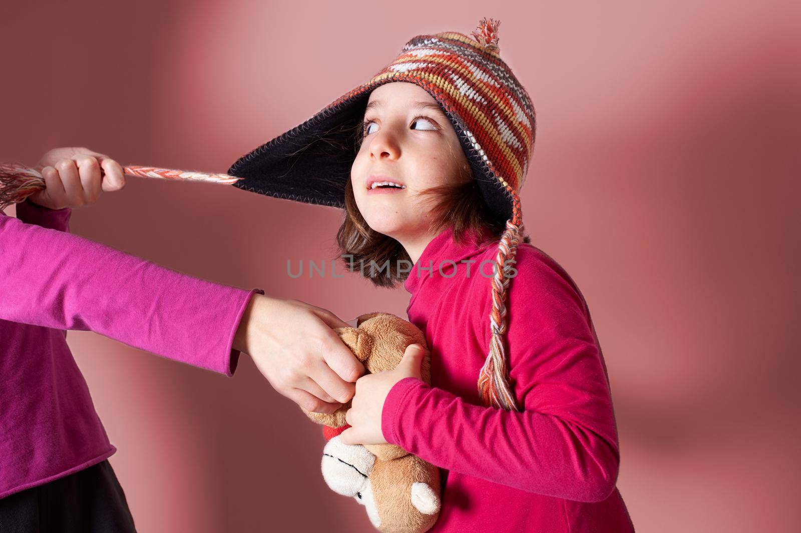 The little sisters who wear winter hats and red pullovers quarrel and fight and compete for a teddy bear. Green bricks background