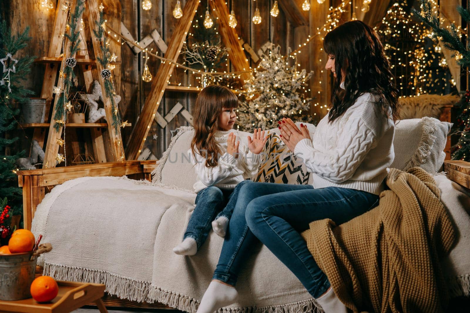 A little girl with her mother in a cozy home environment on the sofa next to the Christmas tree. The theme of New Year holidays and festive interior with garlands and light bulbs.
