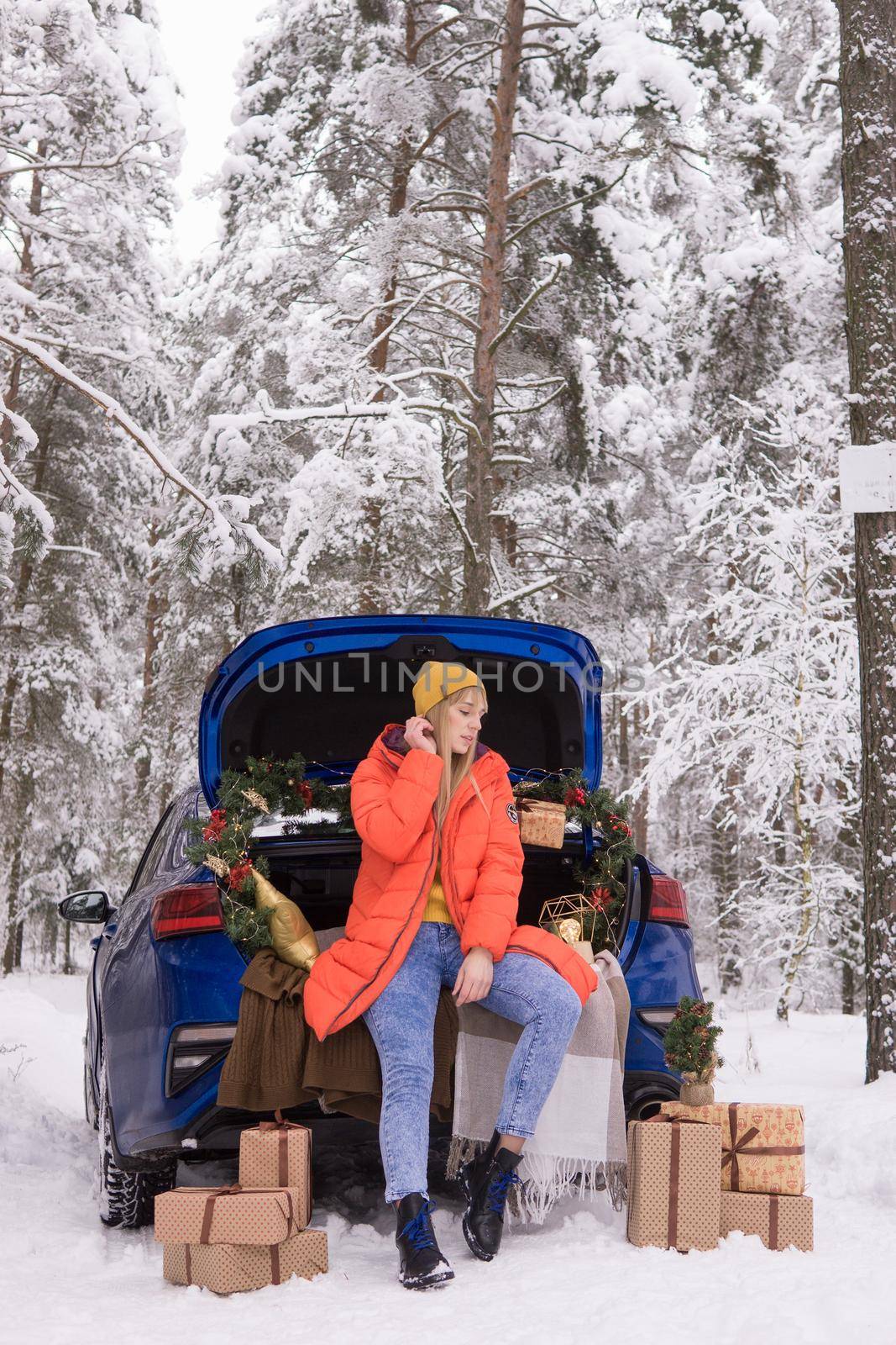 A woman in a winter snow-covered forest in the trunk of a blue car decorated with Christmas decor. The concept of Christmas and winter holidays.