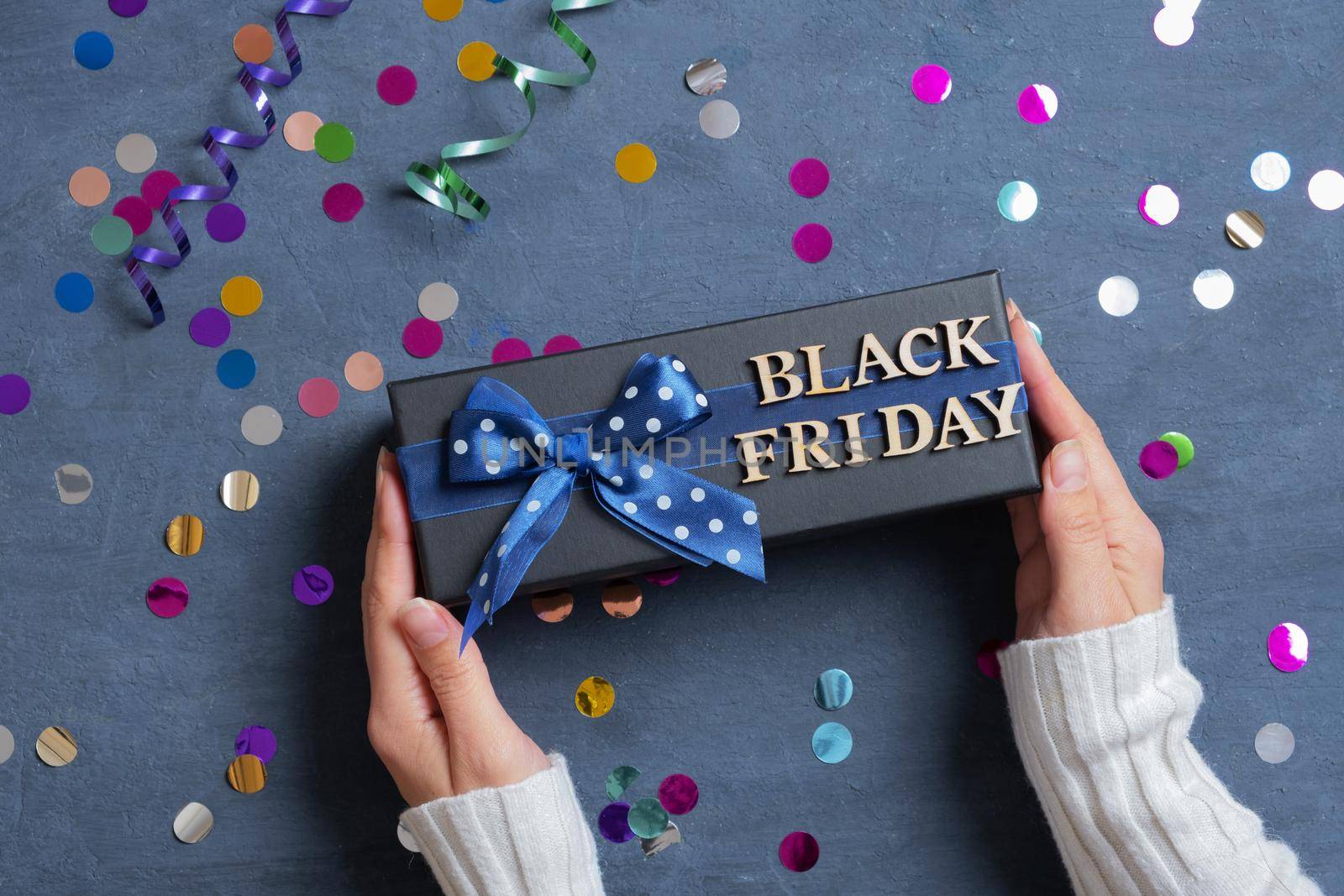 Black Friday text with gift in female hands and festive tinsel flat lay on dark cement background.