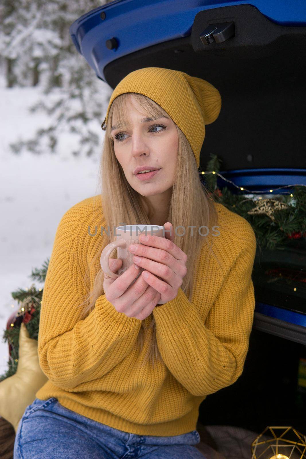 A woman in a winter snow-covered forest in the trunk of a blue car decorated with Christmas decor. The concept of Christmas and winter holidays.