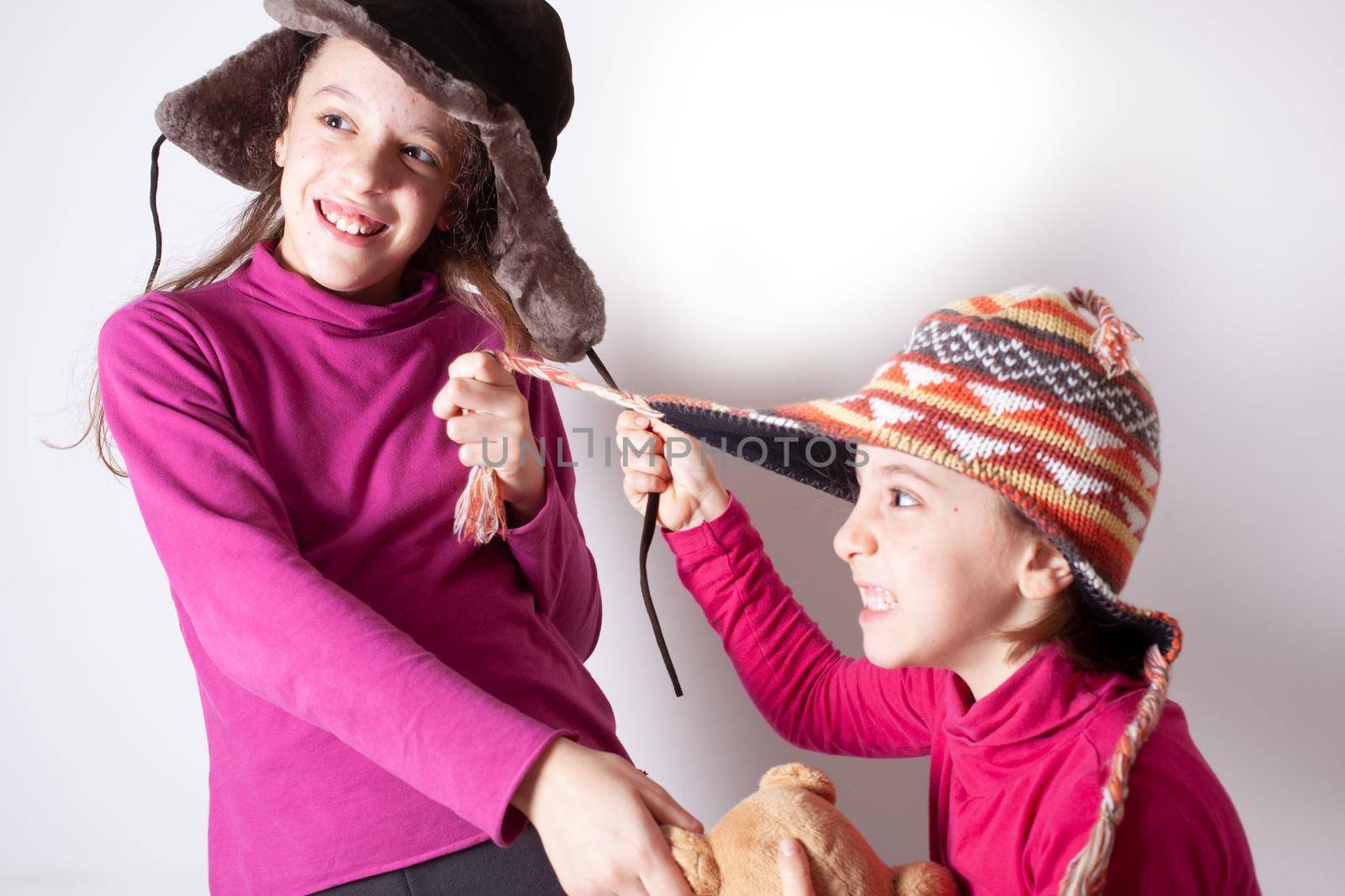 The little sisters who wear winter hats quarrel and fight pulling the laces of hats on white background