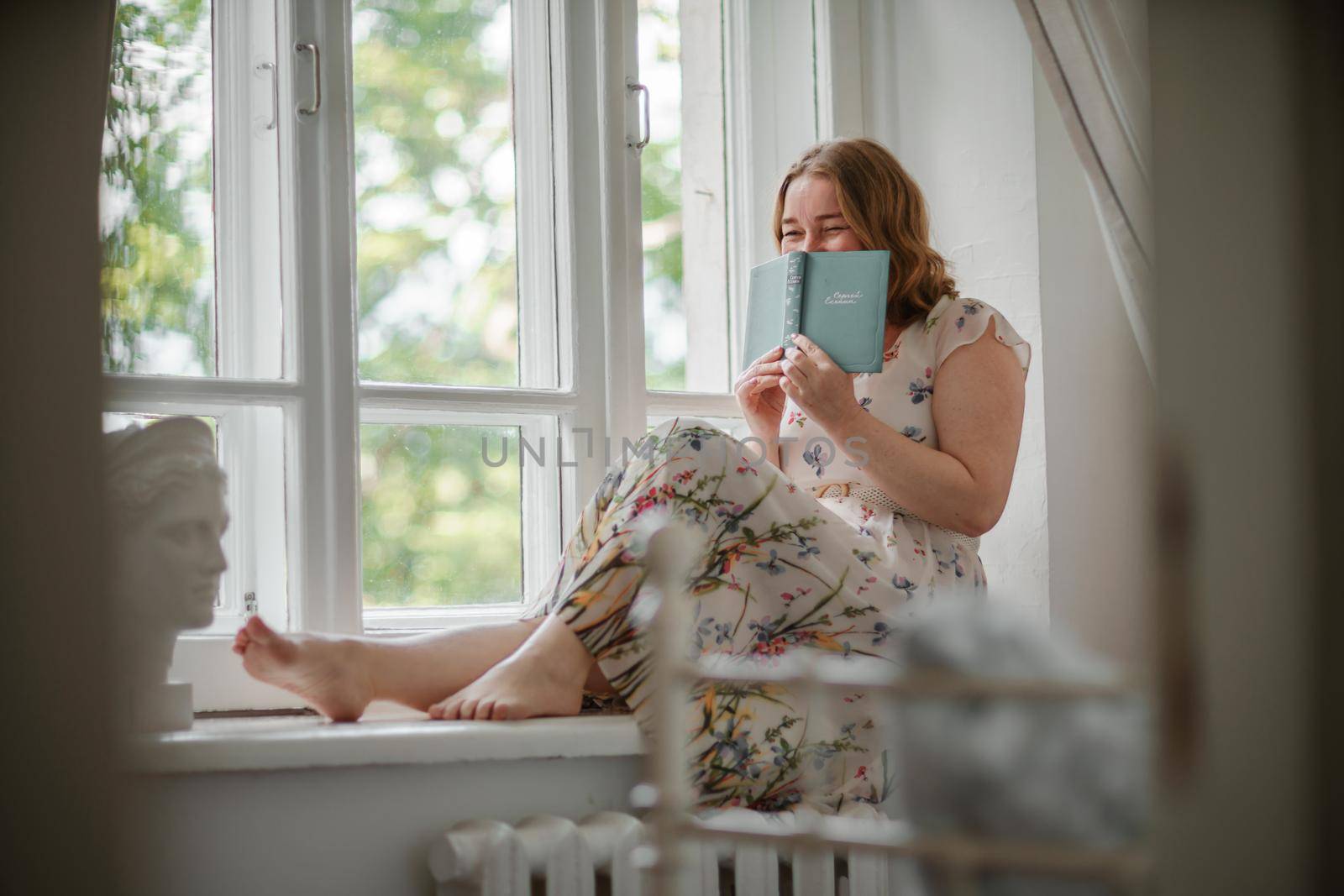 A middle-aged woman in a cream dress sits mysteriously and looks out the window on the windowsill. Green trees outside