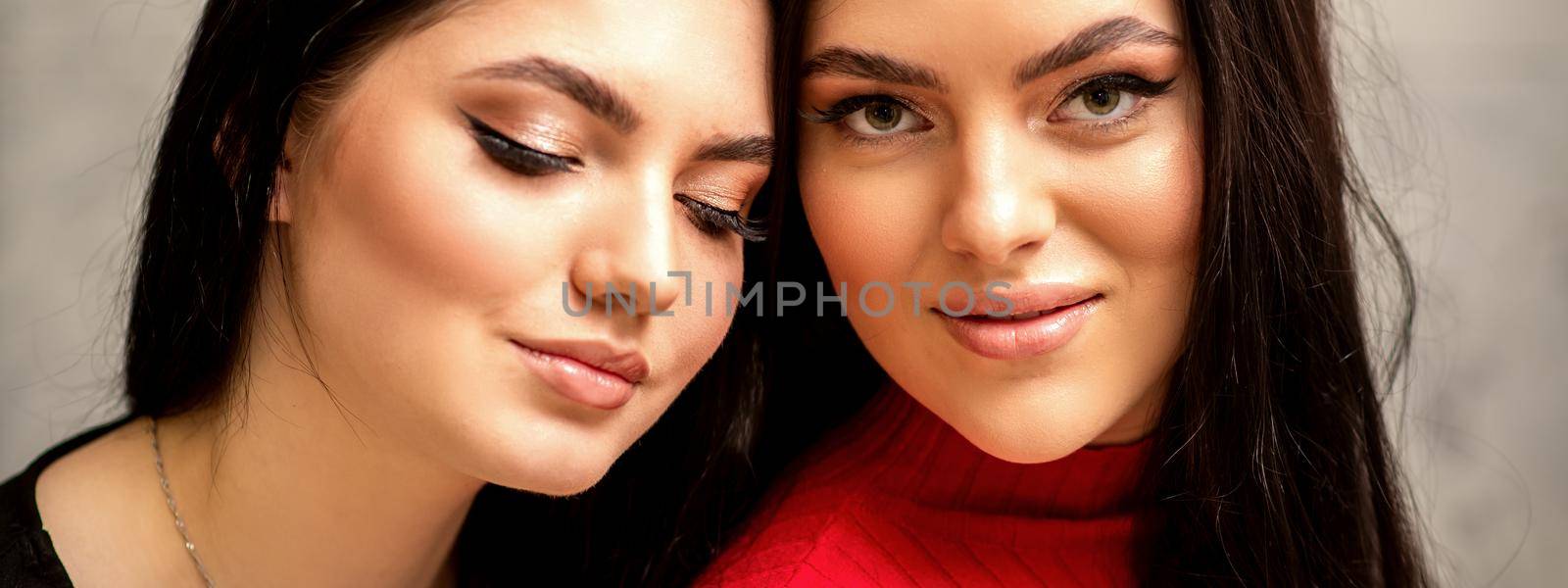Two fashionable young women. Portrait of the two beautiful female models with long hair and makeup. Two beauty young caucasian women with a black hairstyle on the background of a gray wall