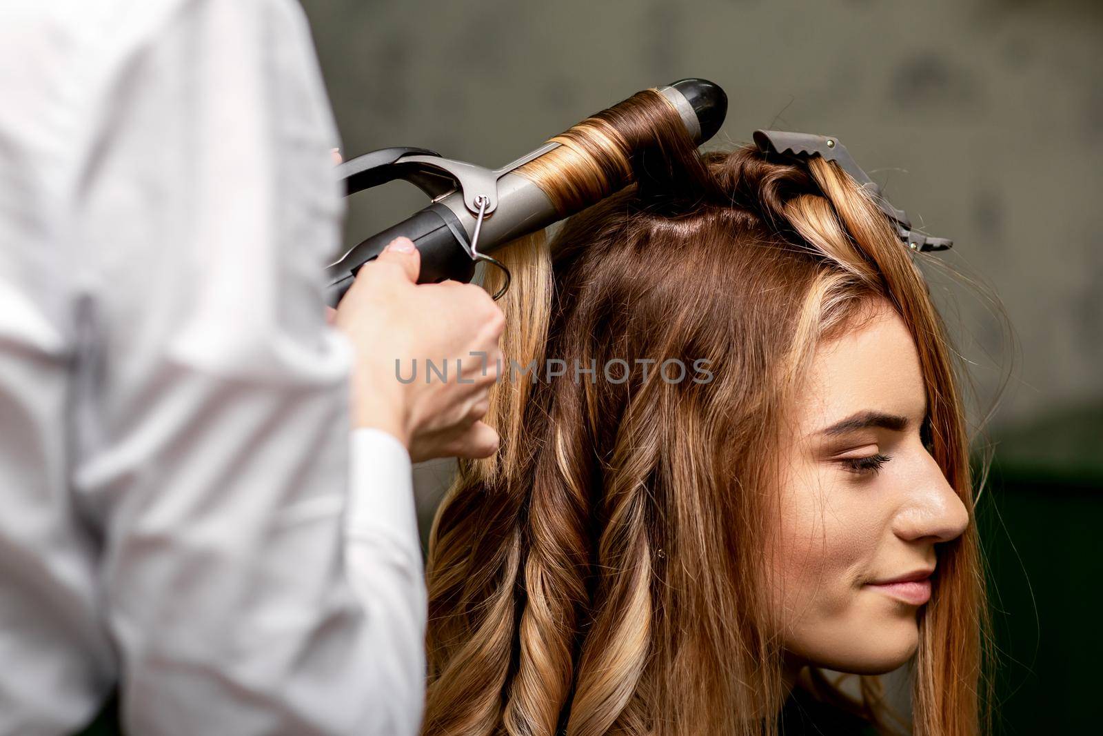 The female hairdresser is curling hair for a brown-haired young caucasian woman in a beauty salon