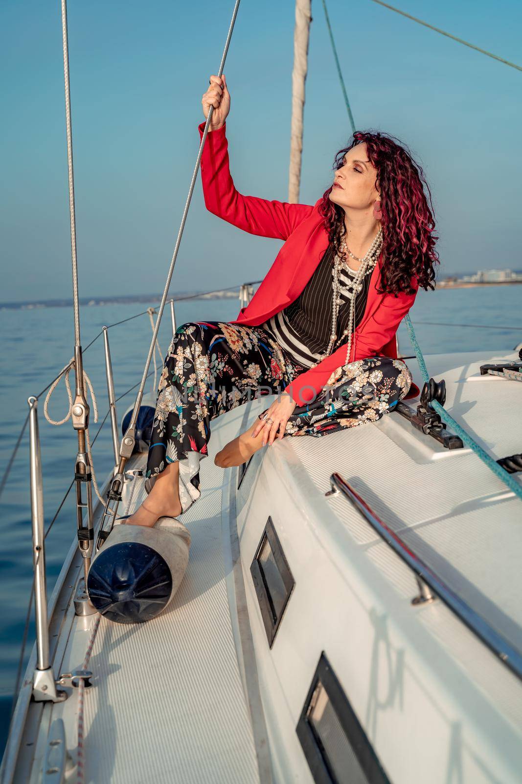 A woman sits on the bow of a yacht on a sunny summer day, the breeze develops her hair, a beautiful sea is in the background.