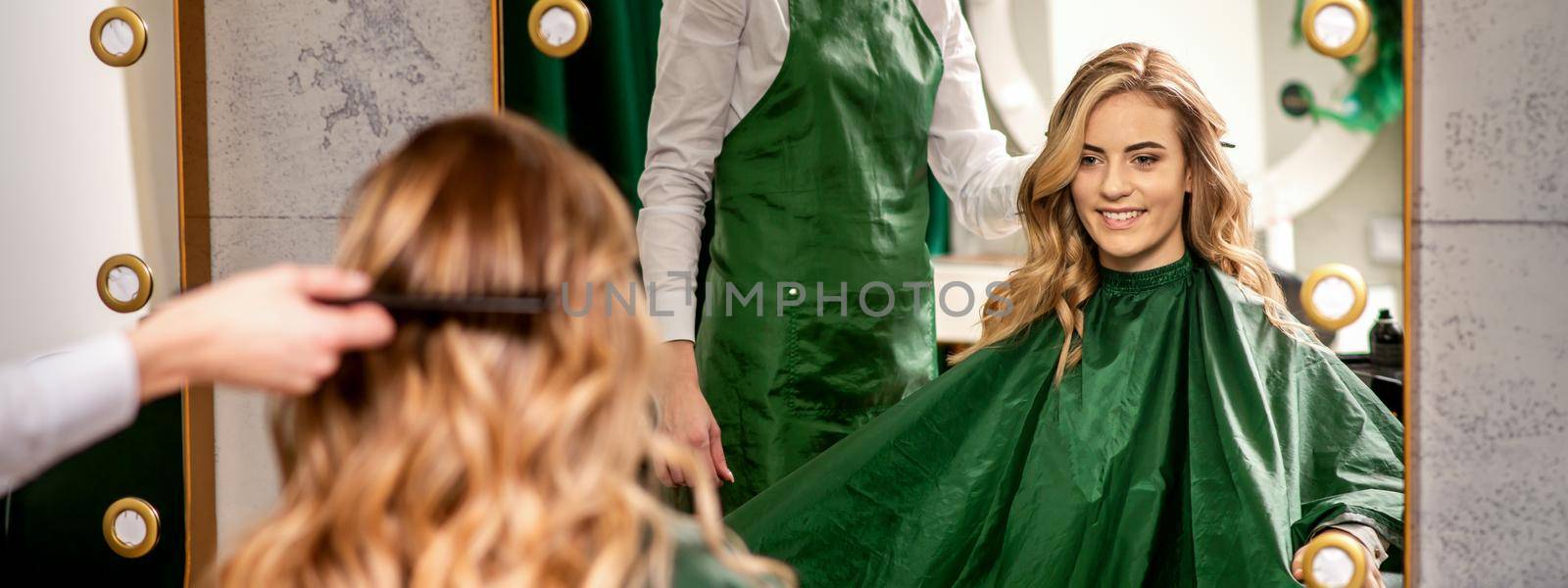 Hairdresser combing long hair of young caucasian woman looking and smiling in the mirror in beauty salon
