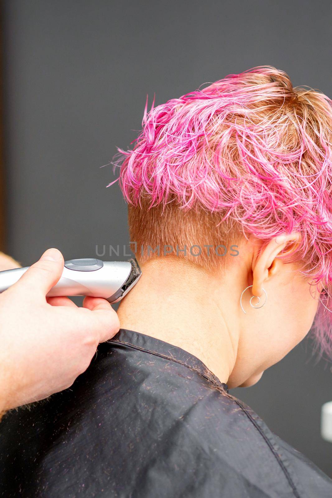 Male hairdresser shaves neck of a young caucasian woman with a short pink hairstyle by electric shaver in a hairdresser salon, close up. by okskukuruza