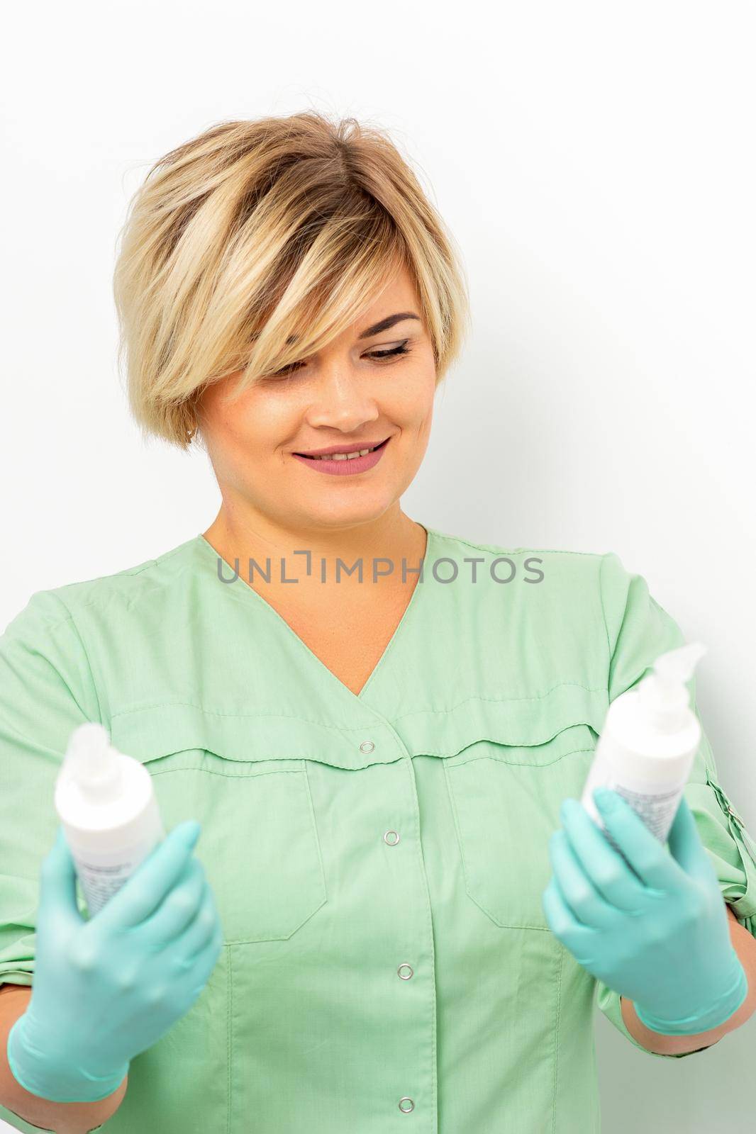 Cosmetics creams and skin care products in the hands of the female beautician smiling and standing over the white wall background. by okskukuruza