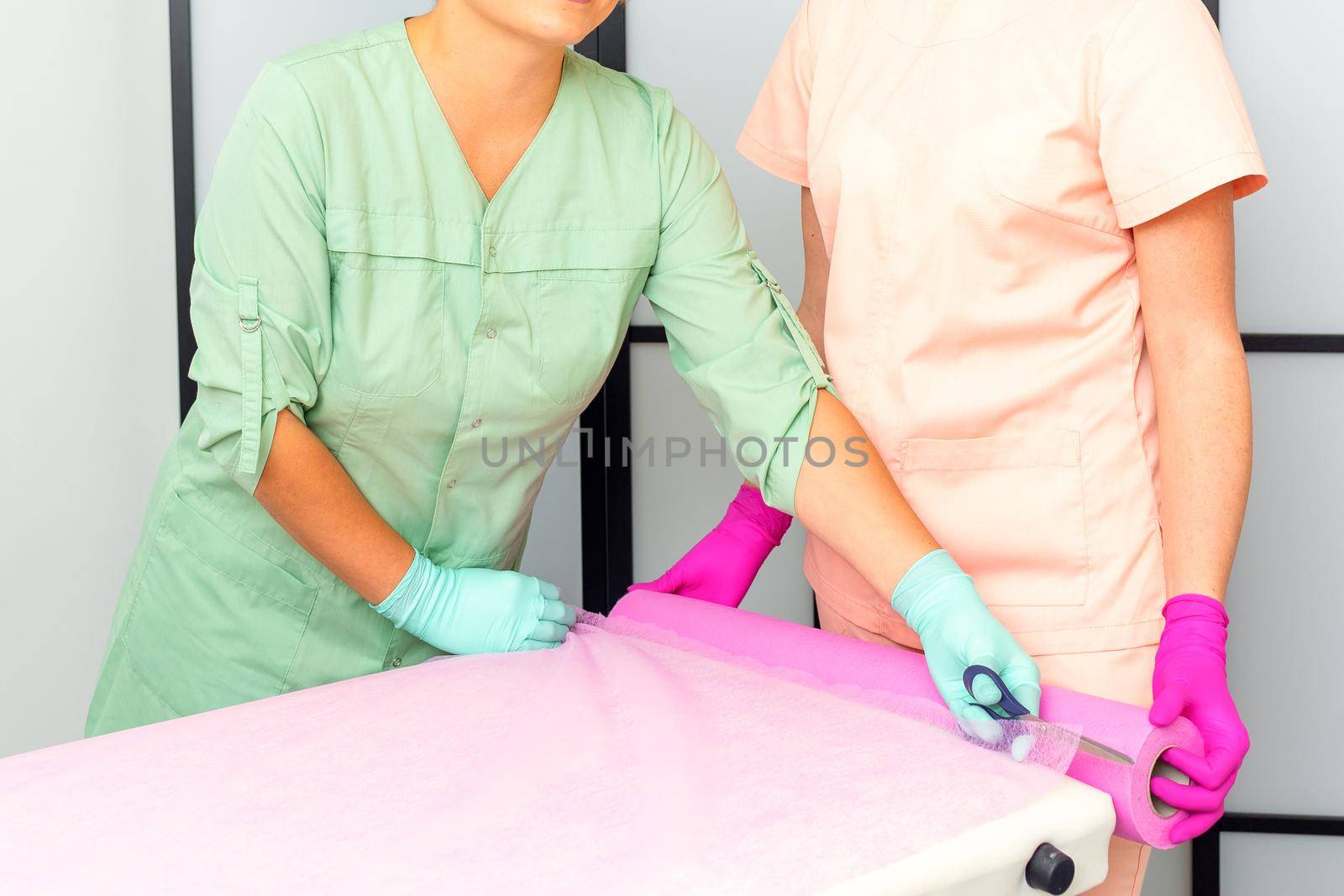 Two medical women cut disposable sheets with scissors during prepare the couch for the beauty procedure. by okskukuruza