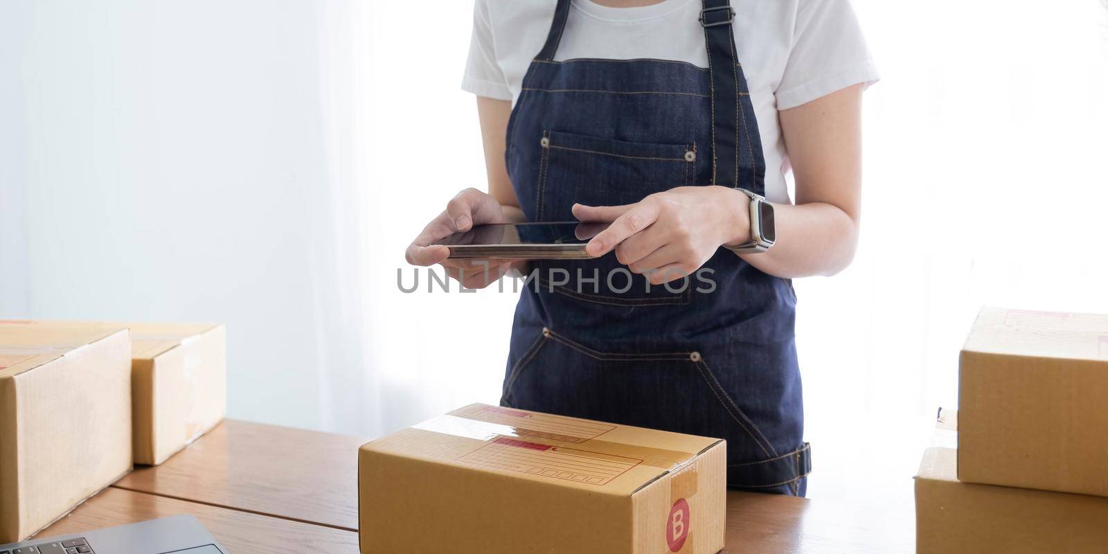 Hands of Asian woman entrepreneur holding smartphone with cardboard boxes, computer laptop, online selling equipments. Packing, business and technology concept. Top view, copy space.