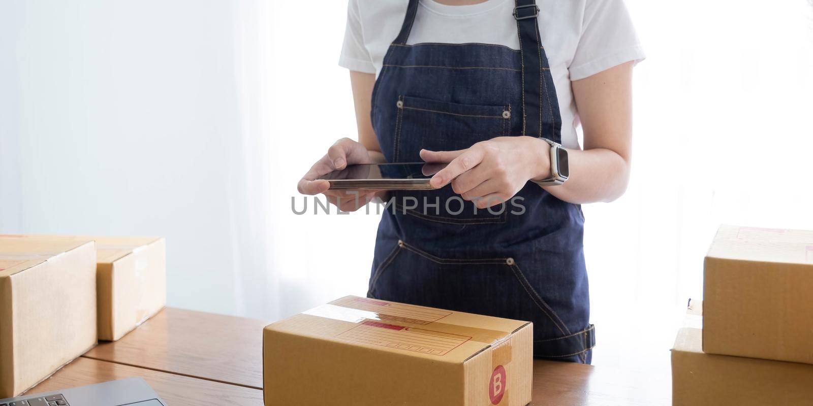 Hands of Asian woman entrepreneur holding smartphone with cardboard boxes, computer laptop, online selling equipments. Packing, business and technology concept. Top view, copy space by wichayada