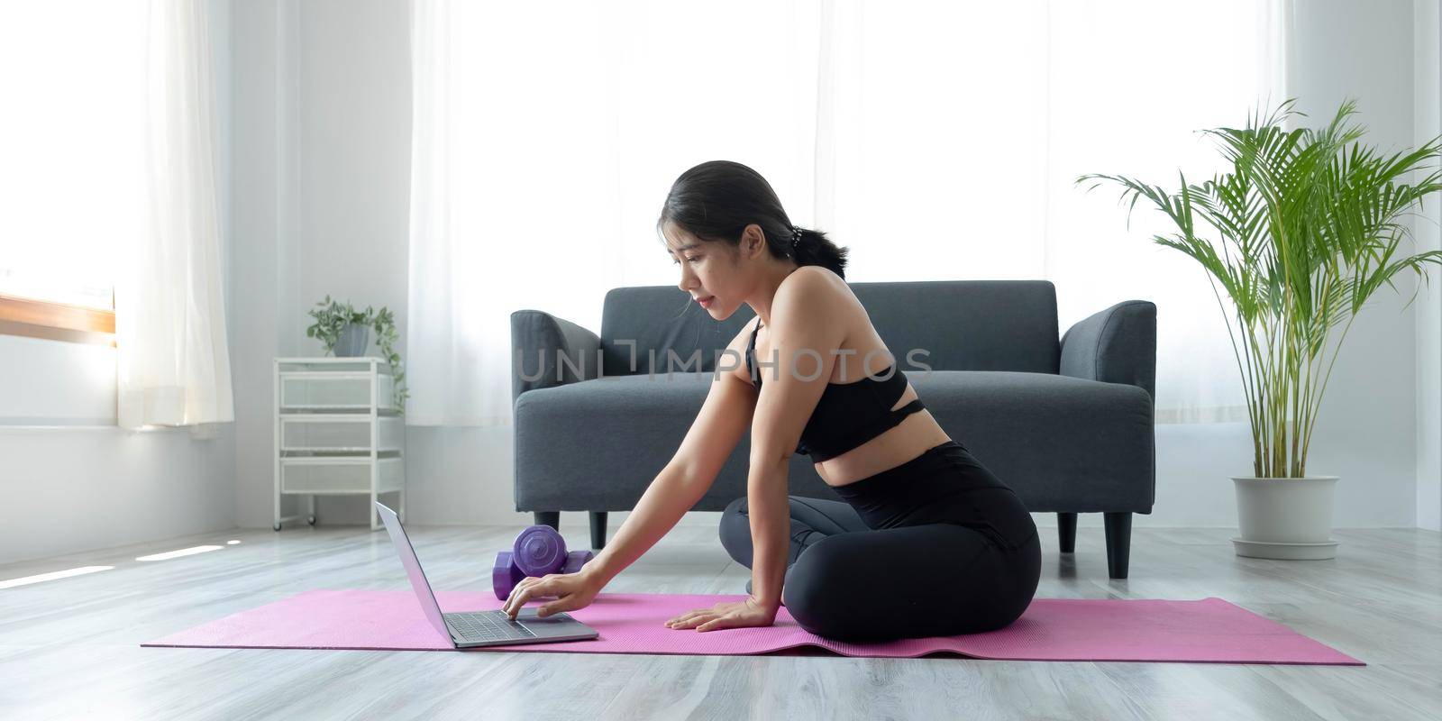 Woman using laptop while exercising at home. Female in sportswear sitting on floor and watching exercise video for her workout. by wichayada