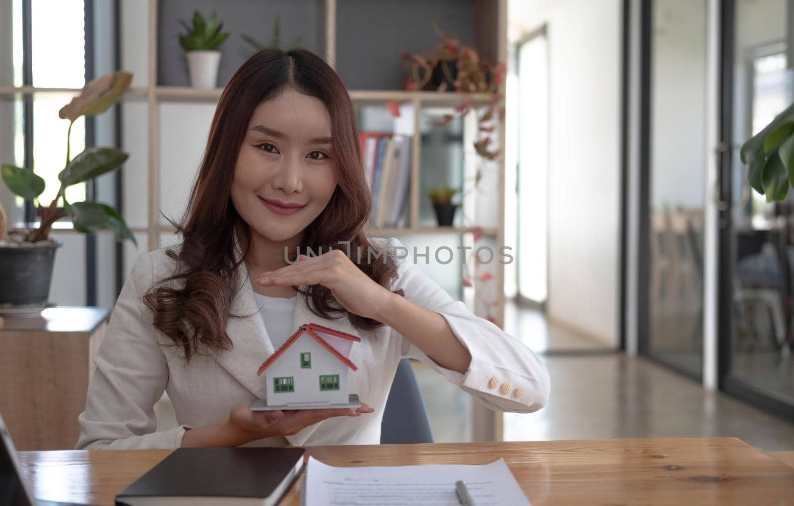 Miniature house in the hands of an Asian woman real estate agent home loan working at the office. Looking at the camera. by wichayada