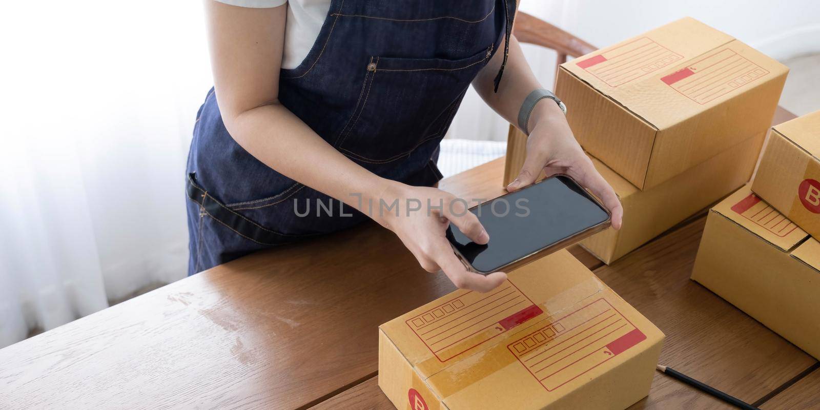 Hands of Asian woman entrepreneur holding smartphone with cardboard boxes, computer laptop, online selling equipments. Packing, business and technology concept. Top view, copy space by wichayada
