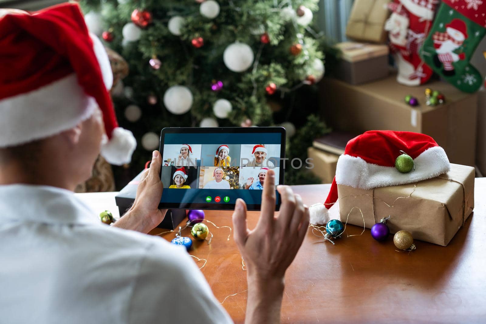 Smiling handsome young man enjoys decorating the Christmas tree while holding a tablet for video call at home. Celebrating the New Year alone