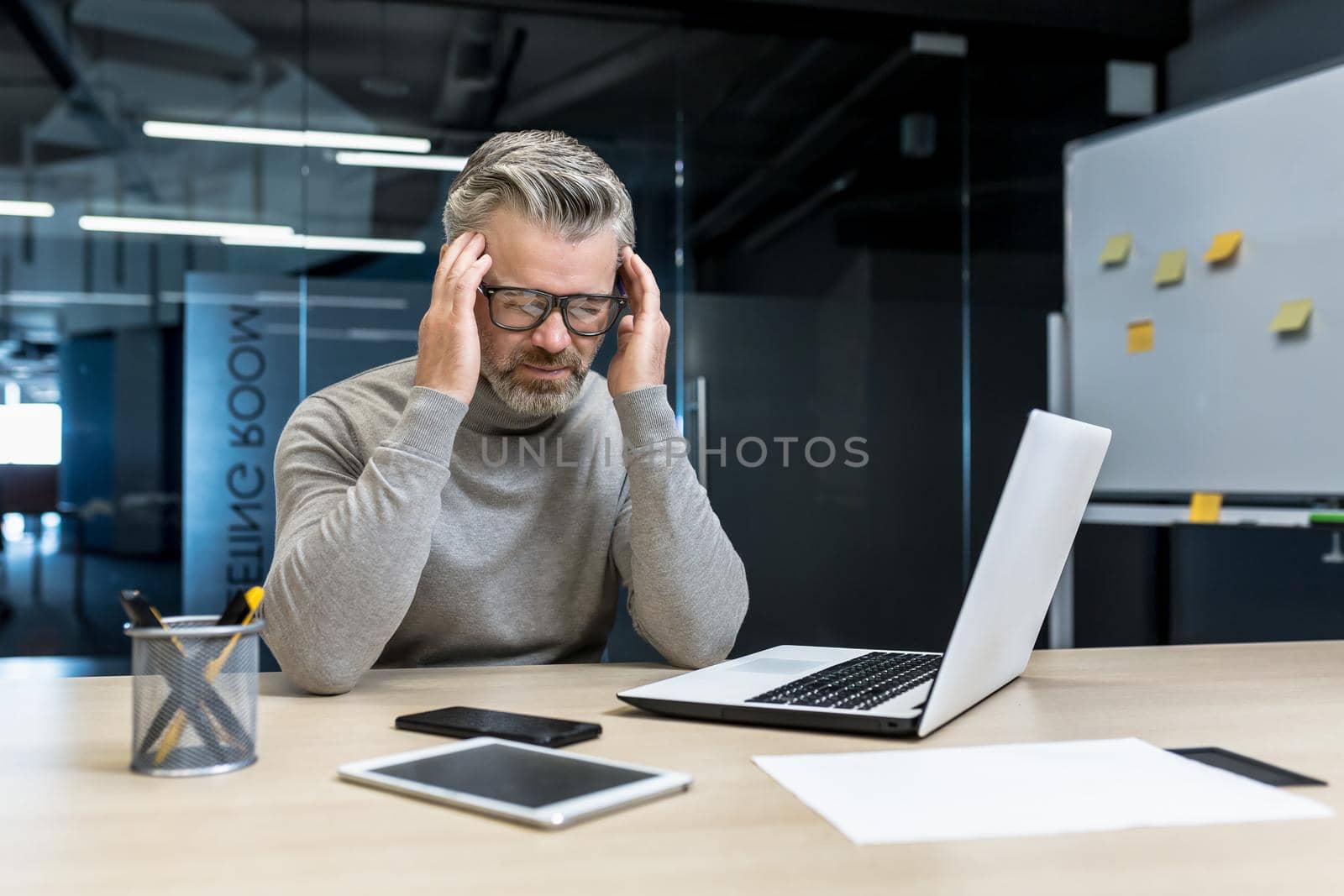 Tension headache at work. A young handsome gray-haired male businessman in glasses sits in the office at a desk with a laptop and a phone, feels a headache, holds his head with his hands.