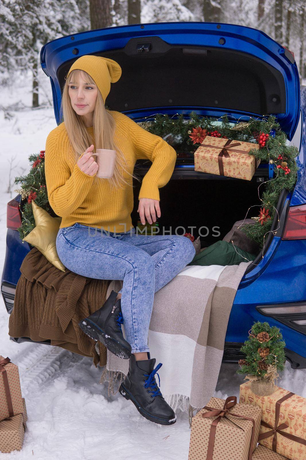 A woman in a winter snow-covered forest in the trunk of a blue car decorated with Christmas decor. The concept of Christmas and winter holidays.