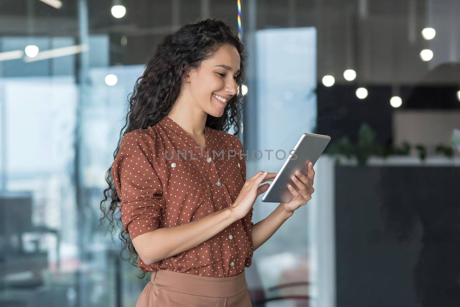 Young beautiful hispanic businesswoman standing in the office in the corridor, using tablet by voronaman