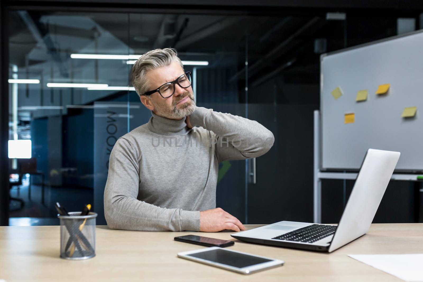 A tired and exhausted businessman man in glasses sits in the office at the desk, holds his neck, massages his neck with his hand. He feels pain in his muscles.