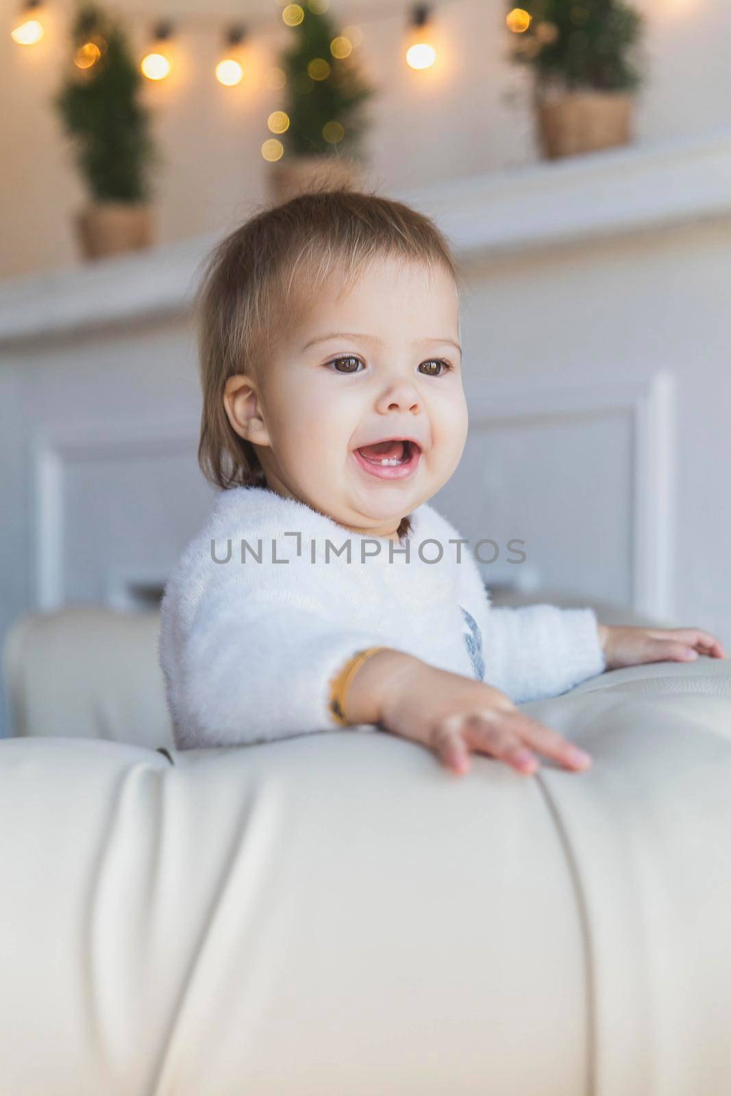 Baby on sofa near christmas tree is smiles so that two teeth are visible.