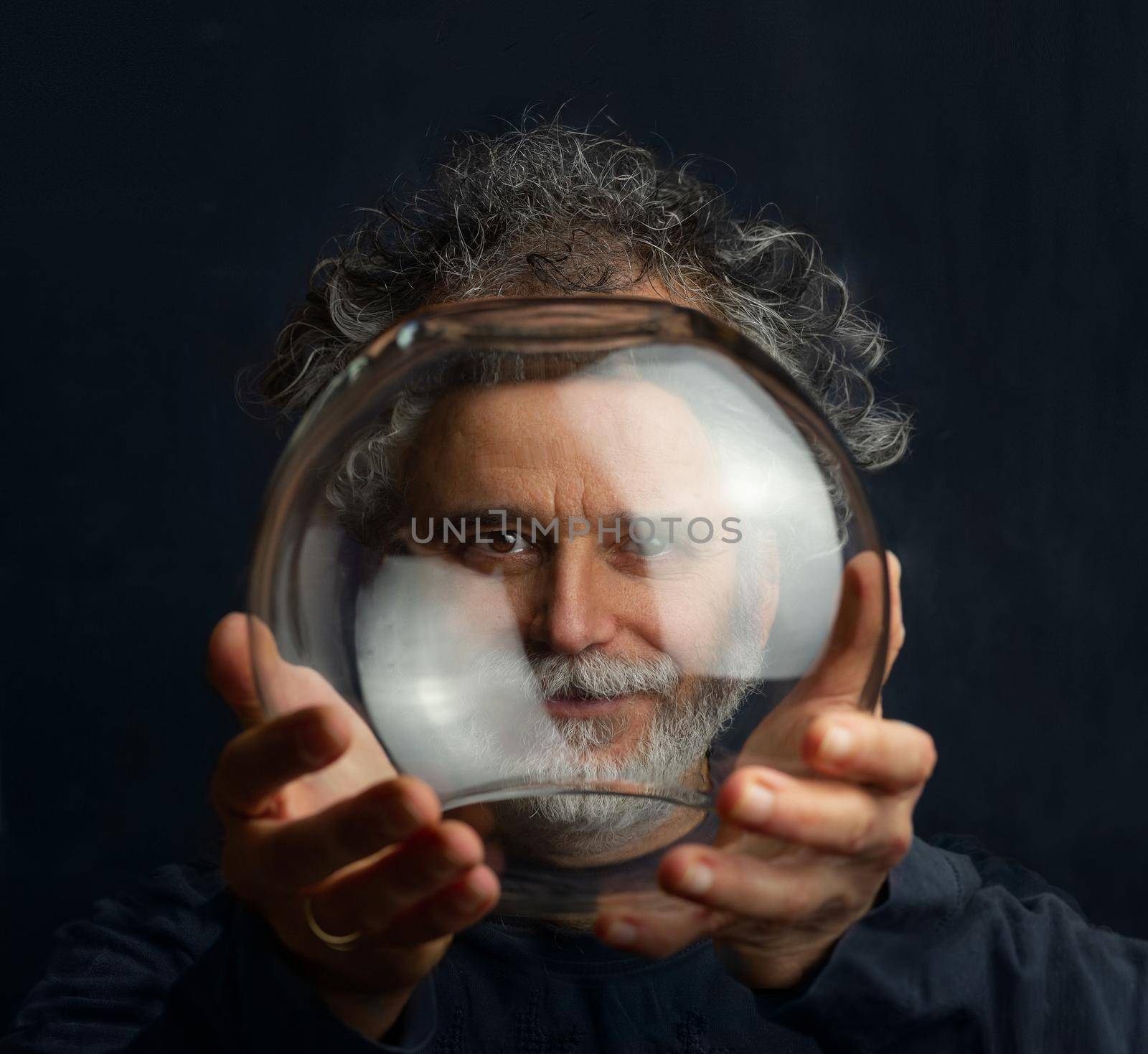 a colorful man with curls, a beard and a silver-colored mustache holds a transparent glass ball in his hands and looks through this ball by Costin