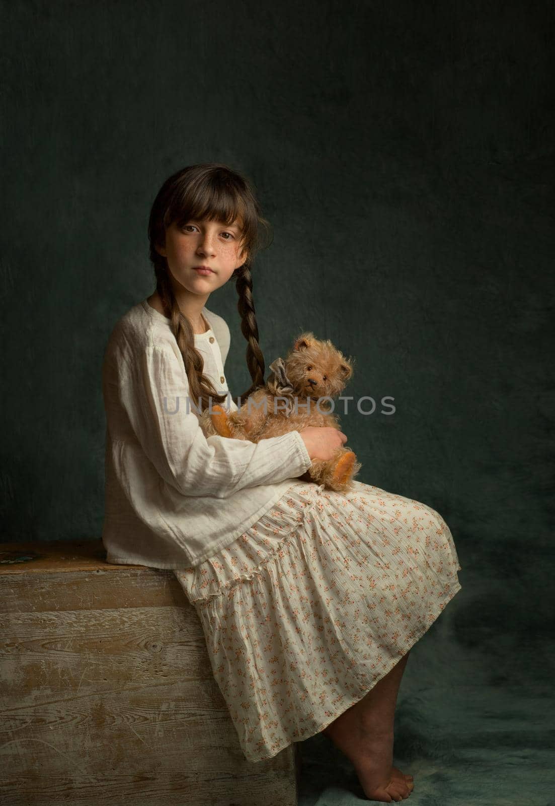a girl with dark hair and two pigtails in a light dress sits with her legs tucked up and holds her favorite toy teddy bear in her hands High quality photo
