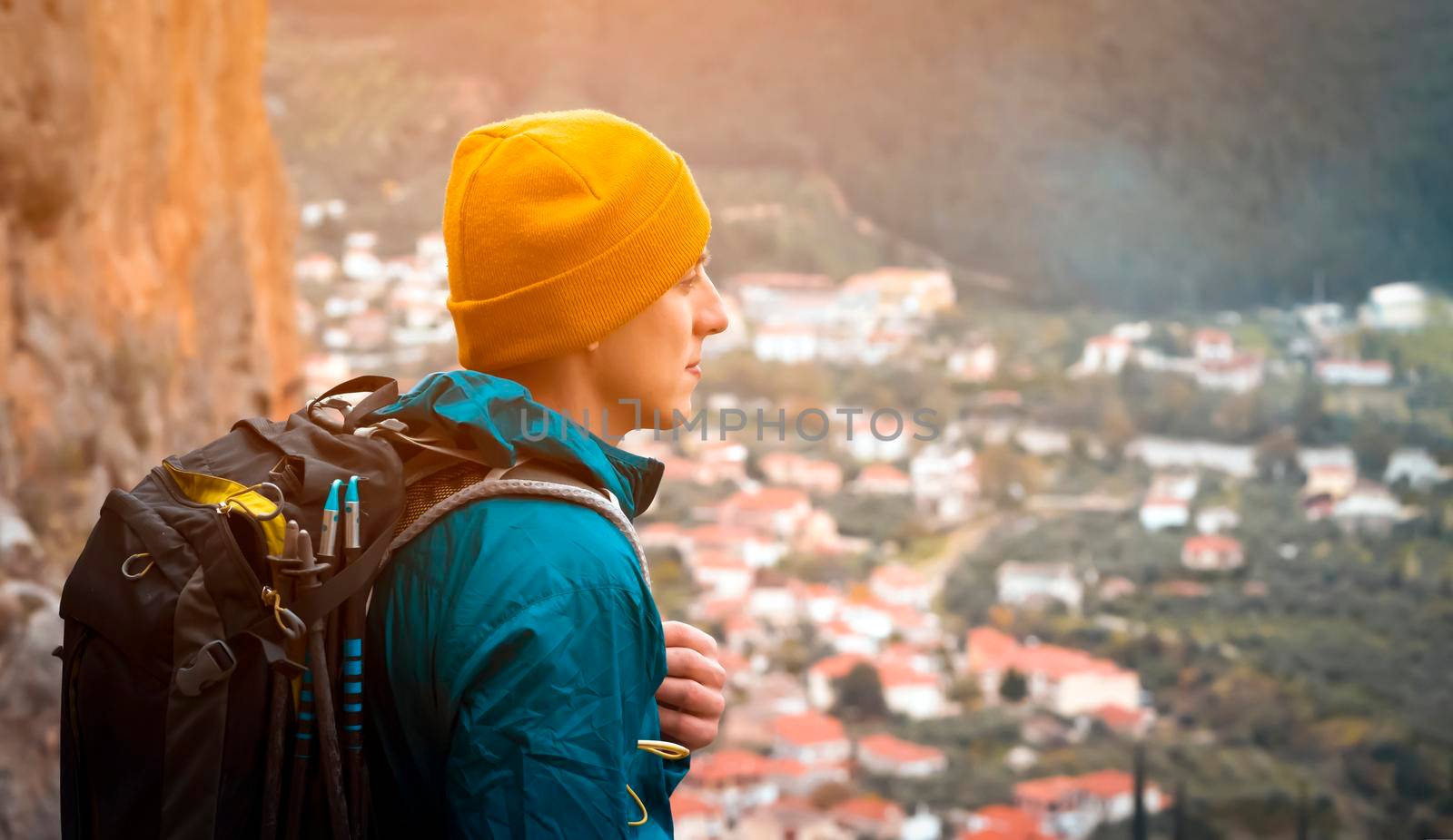 Young man in a yellow hat and with a backpack stands against the background of rocks and a view of a small village with red roofs, is engaged in trekking, discovers the beauty of the Mediterranean.