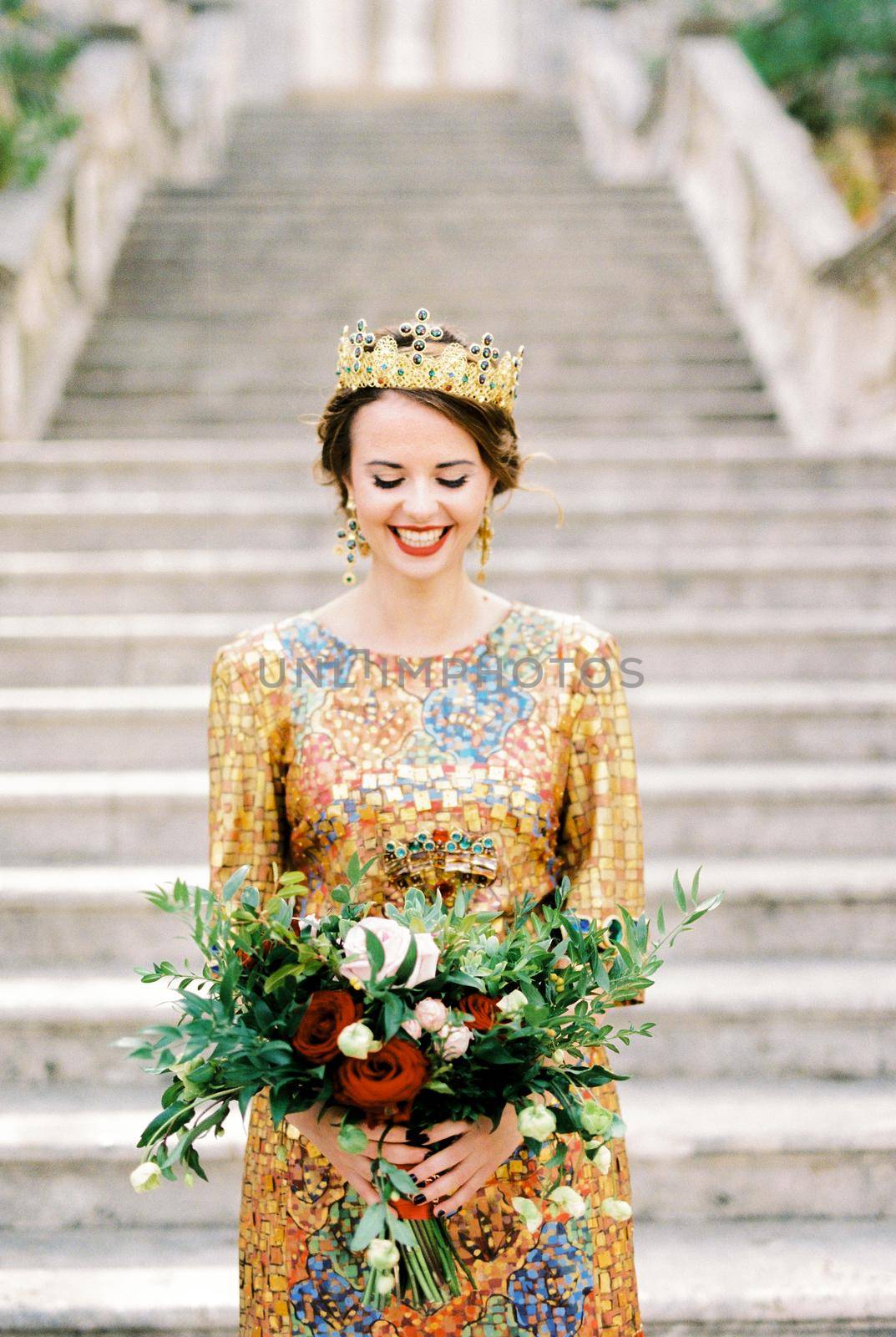 Smiling bride in a golden dress and crown stands on the steps with a bouquet. High quality photo