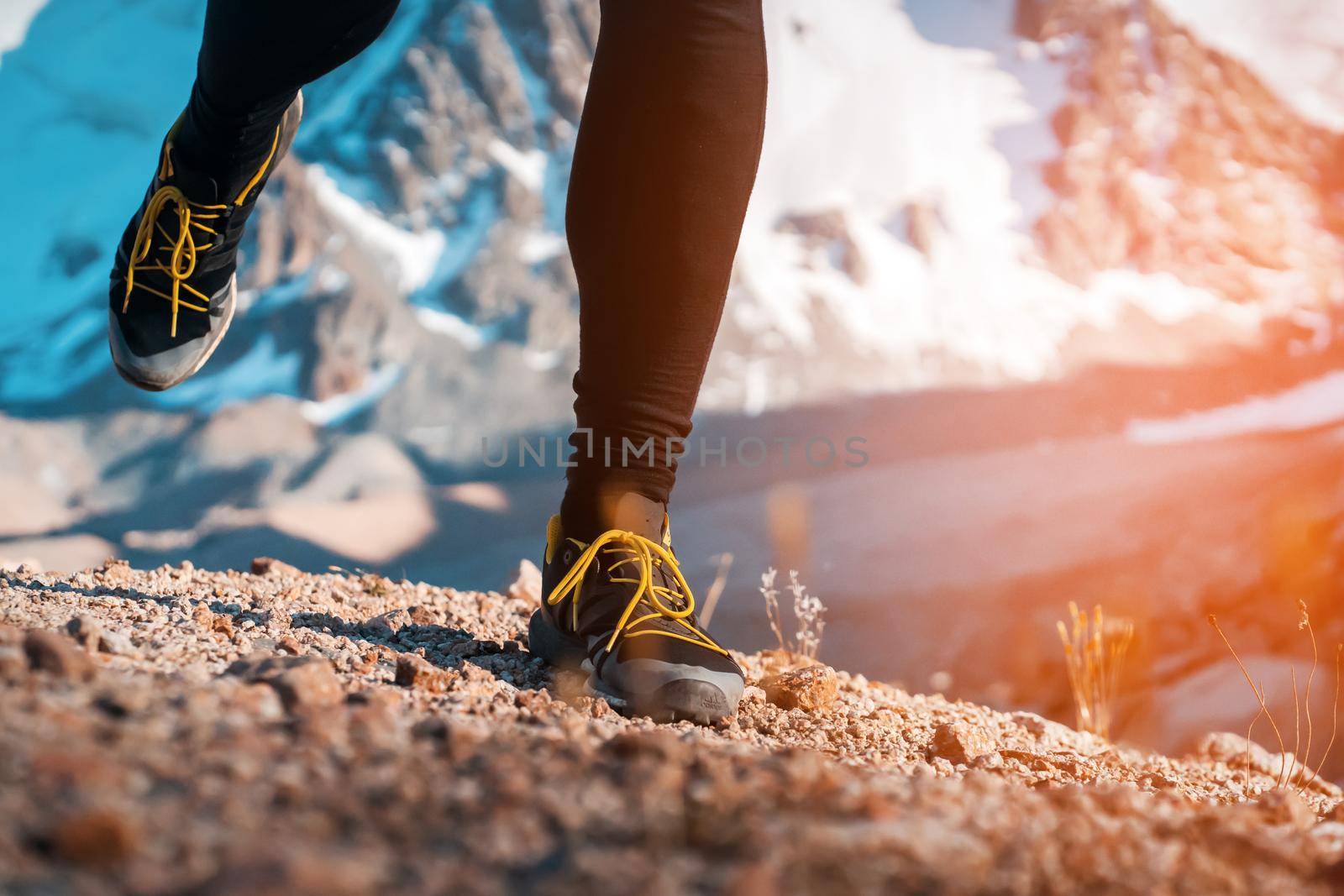 Athlete runner legs run on a trail along a rocky path close-up. Fitness and warm-up, young man with snow-covered mountain peaks in the background and open space around him at sunset.