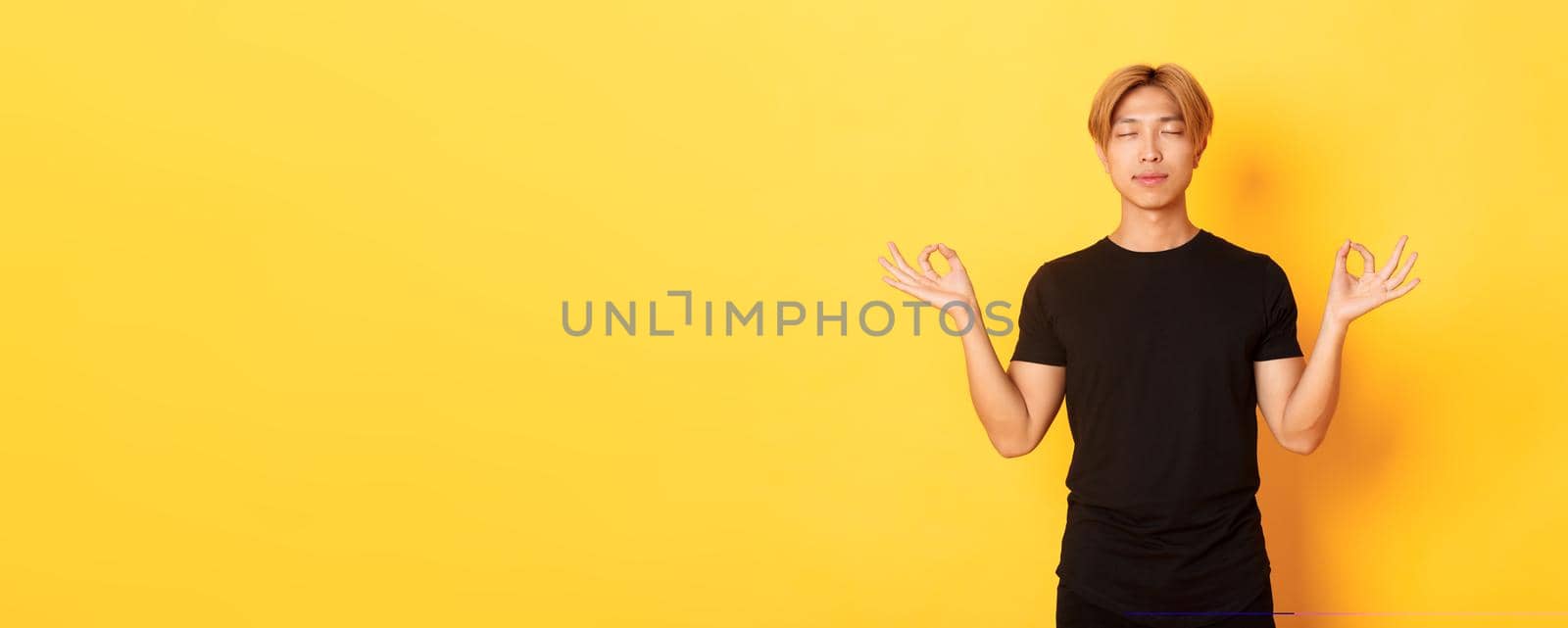 Portrait of relaxed and peaceful handsome asian man with blond hair, close eyes and meditating, attend yoga, standing yellow background by Benzoix