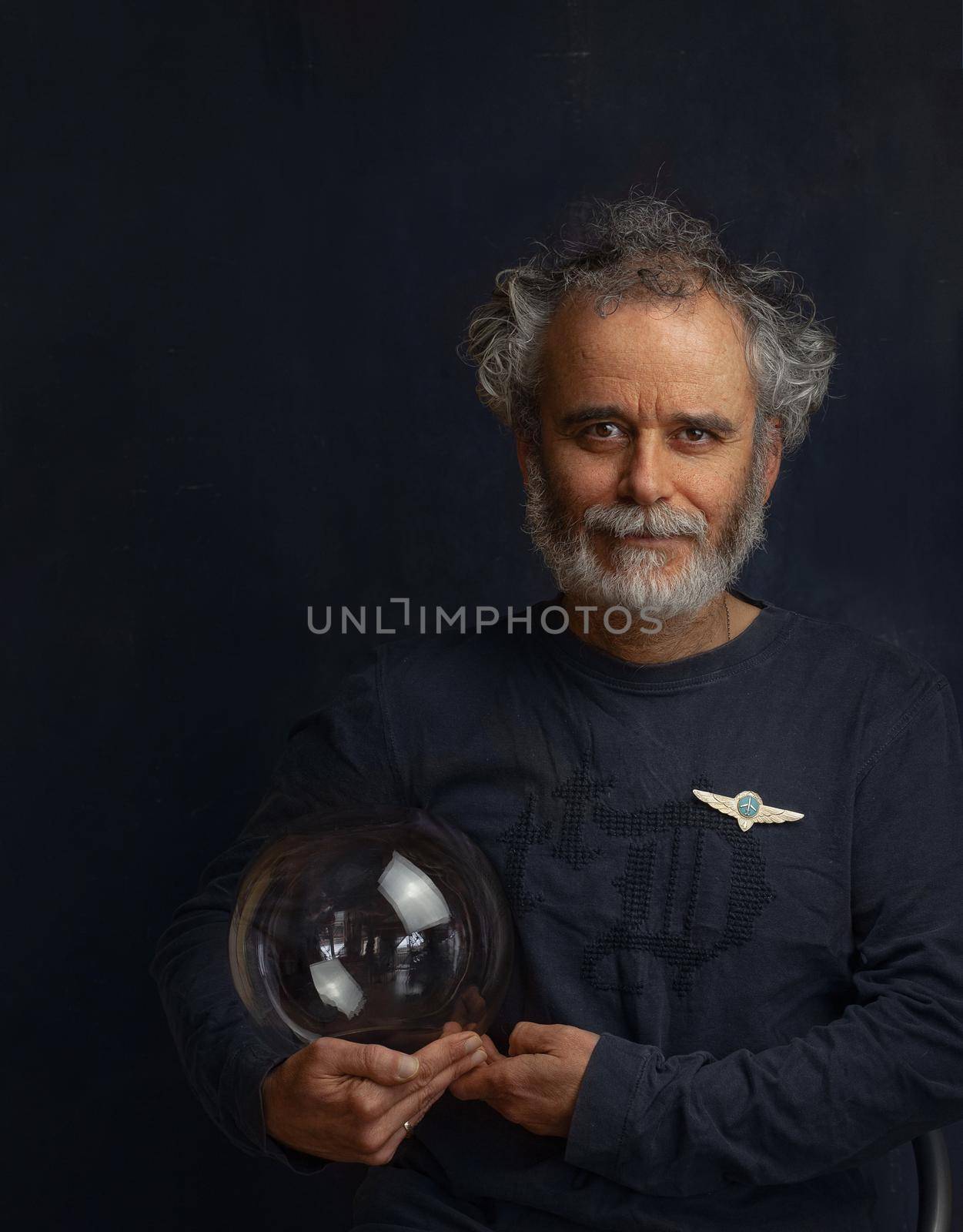 a colorful man with a curled beard and a silvery mustache holds a transparent glass ball in his hand and looks forward smiling a little on his chest civil aviation badge High quality photo