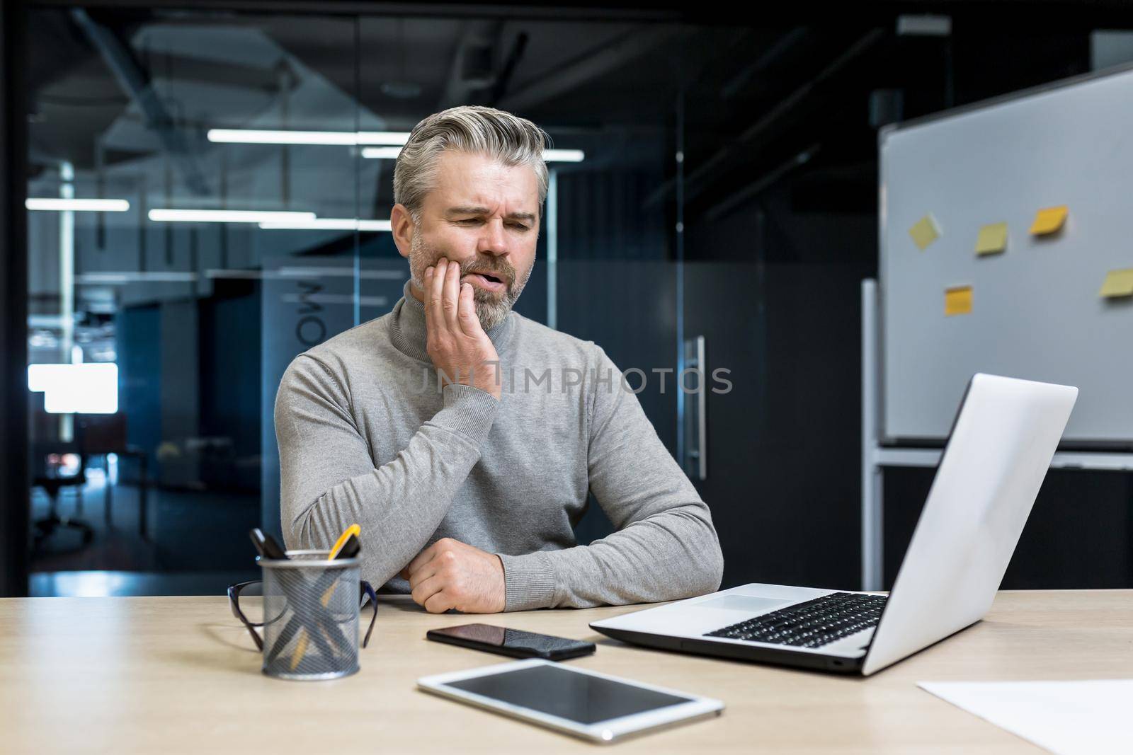 Toothache at work. Handsome gray-haired man, office worker, freelancer sits at a desk with a laptop and a phone, holds his cheek. He feels a strong toothache, needs medical help.