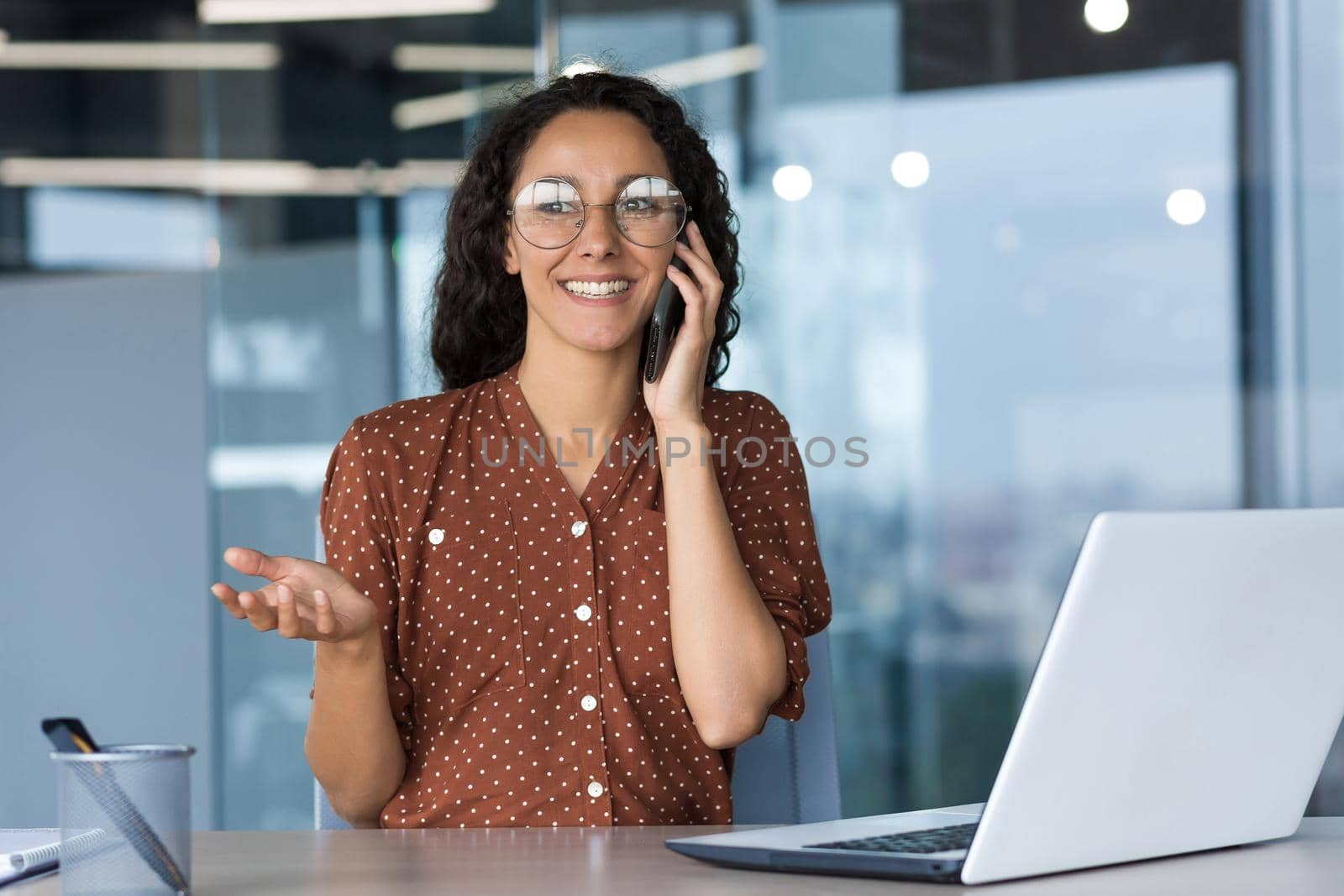 Young beautiful hispanic female freelancer wearing glasses sitting in office at desk with white laptop and talking on phone. He smiles, gestures with his hands.